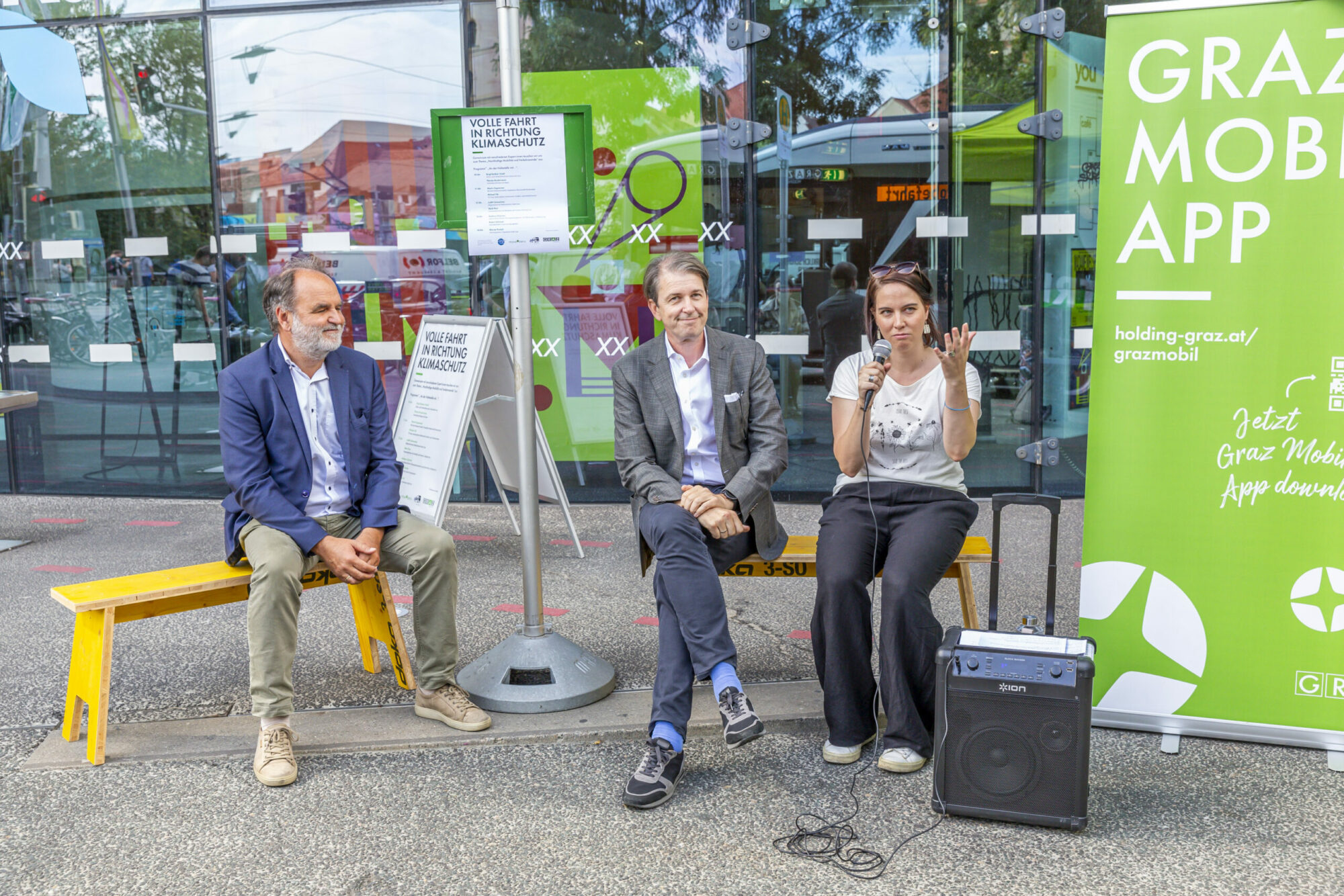 Drei Personen sitzen bei Diskussion vor Banner der GrazMobil-App, Mikrofon und Lautsprecher dabei.