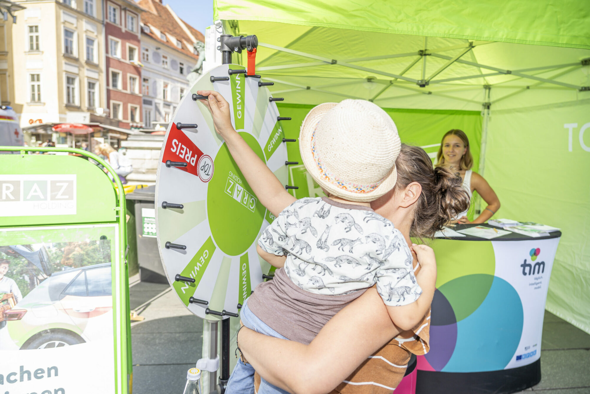 Frau mit Kind dreht ein Glücksrad an grünem Stand der Holding Graz bei sonnigem Wetter.