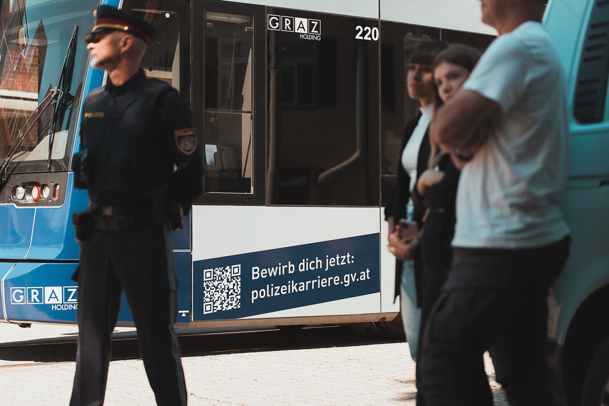 Polizist vor Straßenbahn mit Werbung für Polizeikarrieren und QR-Code, Menschen im Hintergrund.