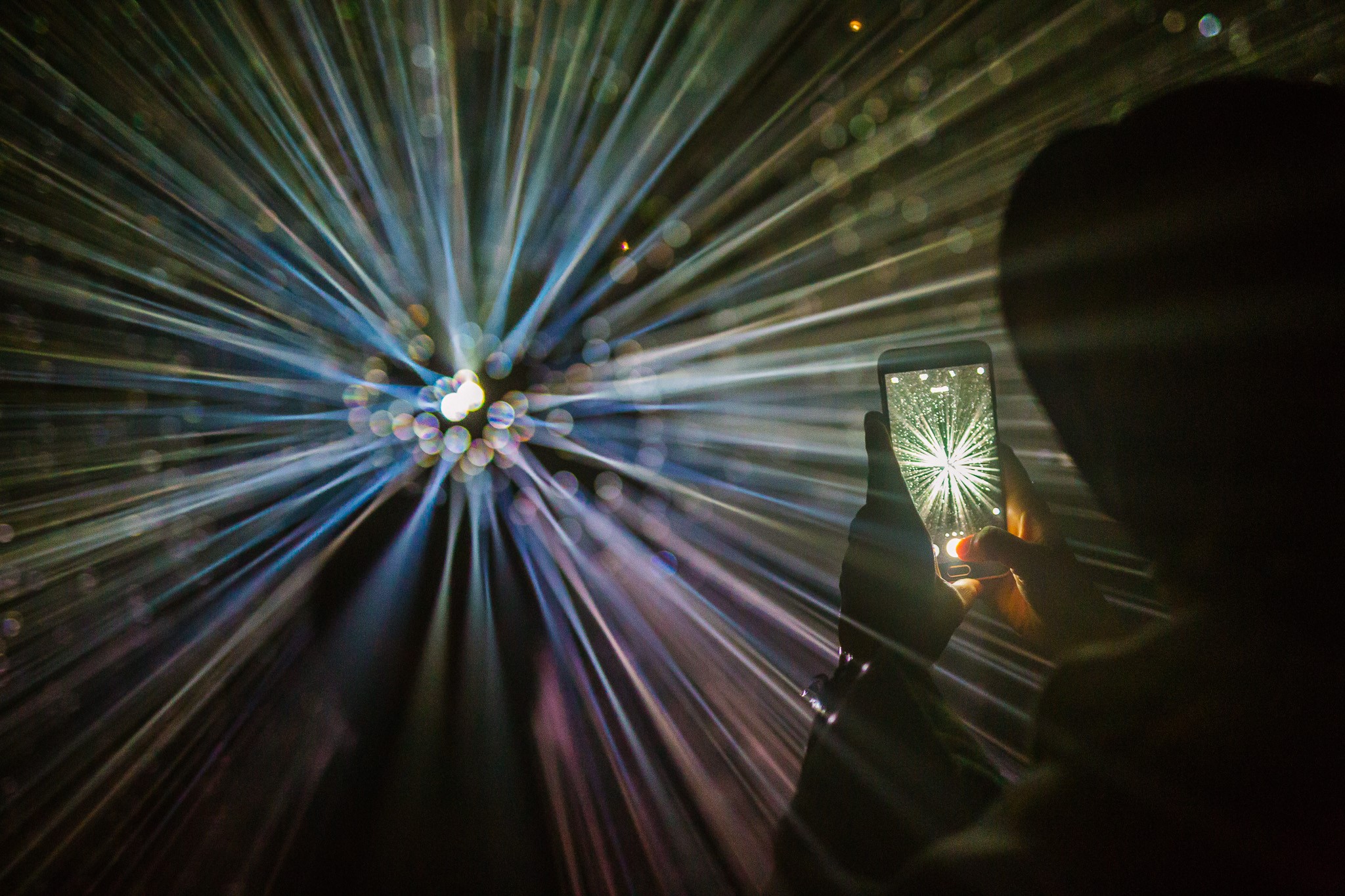 Person fotografiert ein Lichtkunstwerk mit einem Handy, das strahlenförmige Lichteffekte zeigt.