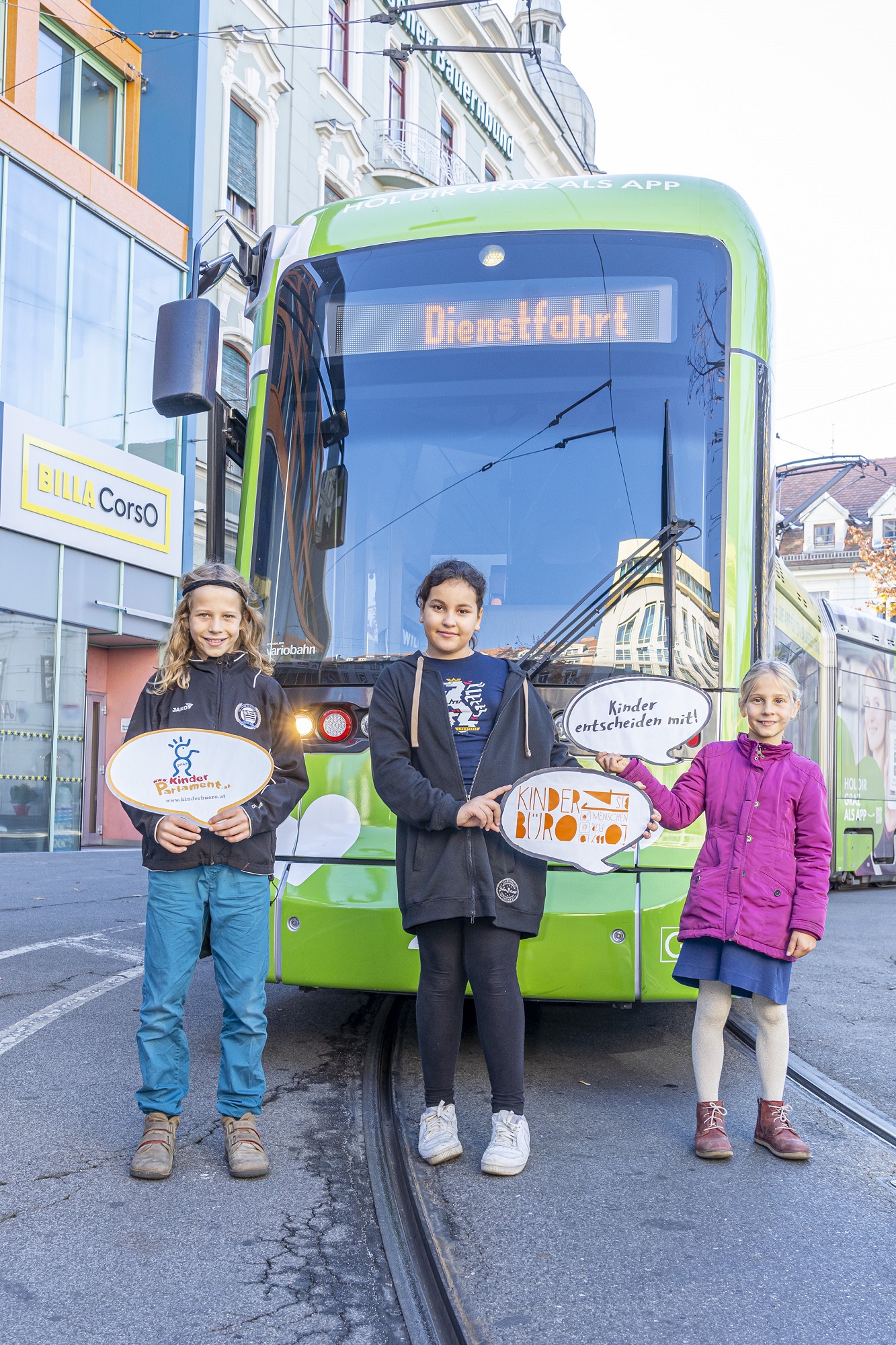 Drei Kinder stehen vor einer grünen Straßenbahn und halten Schilder für Kinderrechte in den Händen.