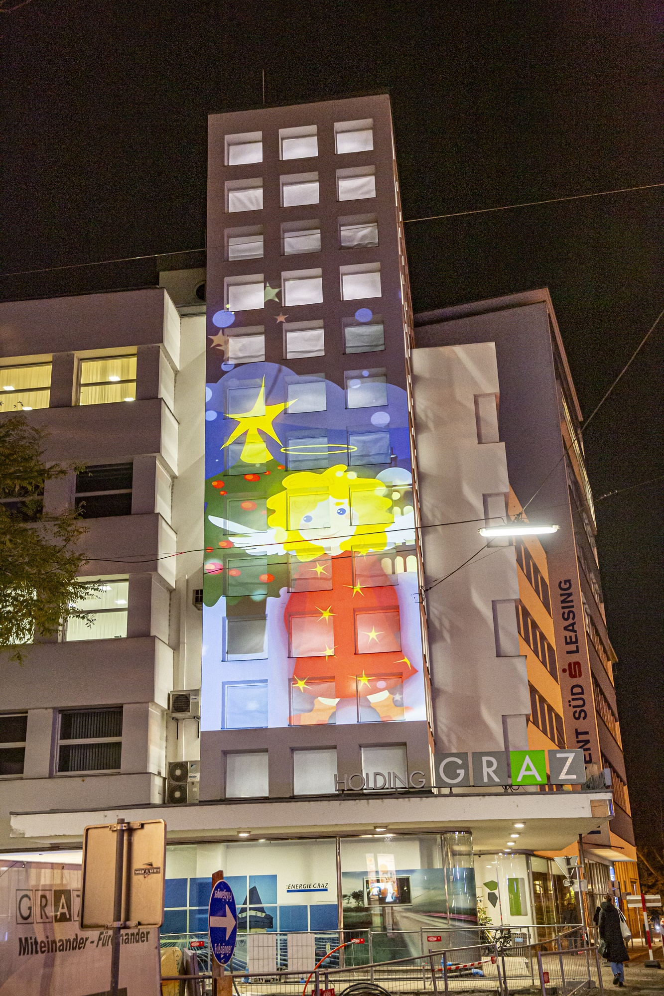 Holding Graz-Gebäude bei Nacht mit großer, bunter Weihnachtsengel-Projektion auf Fassade.