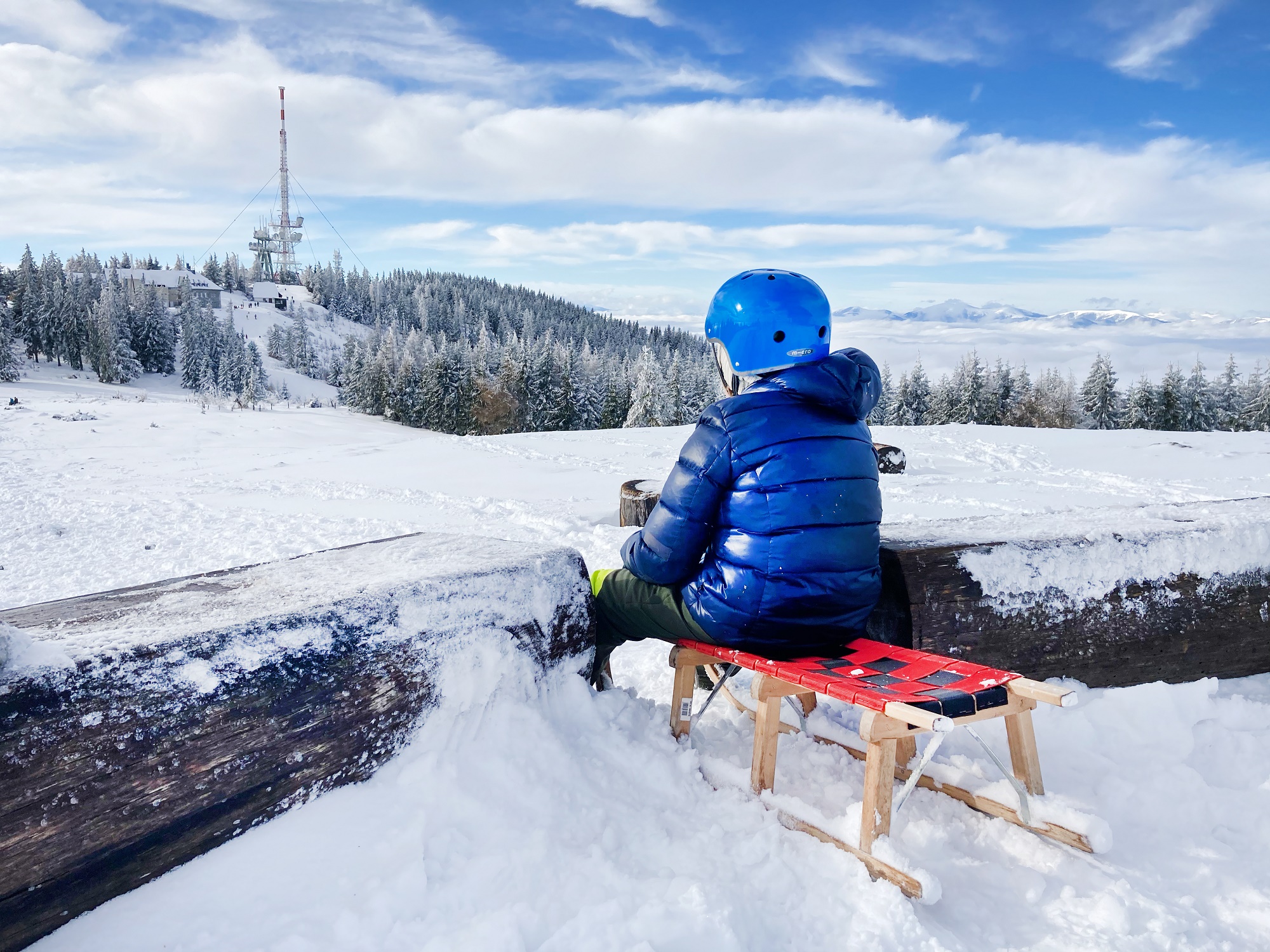 Kind in blauer Winterjacke und Helm sitzt auf einem Schlitten im Schnee, mit Blick auf Schöckl-Gipfel.