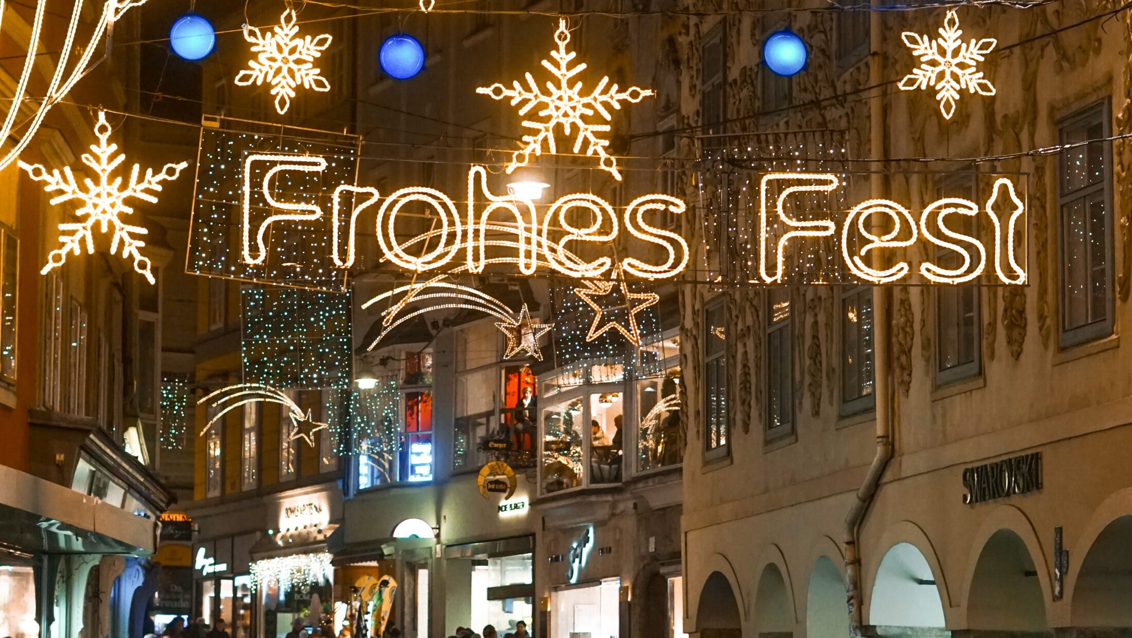 A crowd of people walks down a street in Graz, Austria, decorated with Christmas lights and the sign "Frohes Fest" (Happy Holidays).