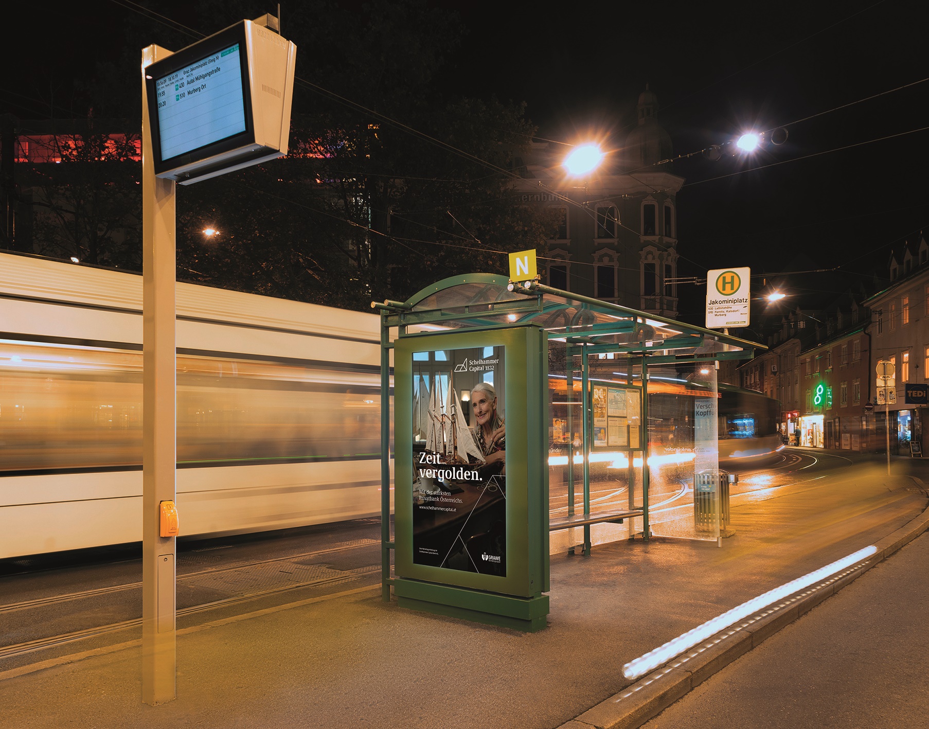 Haltestelle in Graz bei Nacht mit leuchtendem Werbeplakat und vorbeifahrender Straßenbahn.