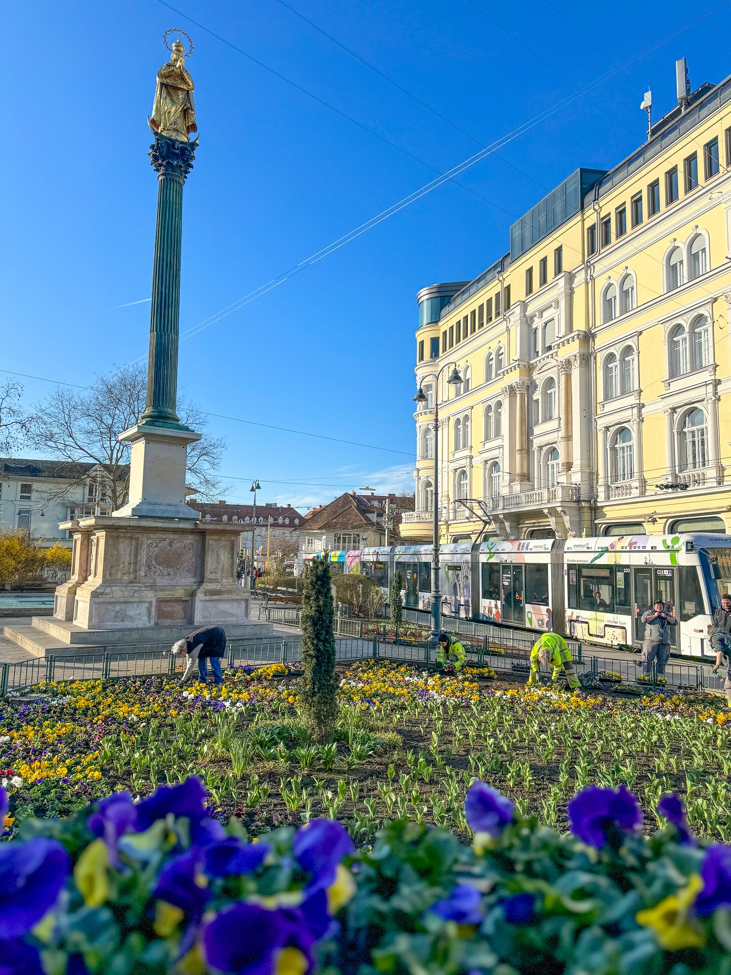 Blick auf das Eisernen Tor in Graz mit einer Statue auf einer Säule und Gärtnern, die ein Blumenbeet bepflanzen.