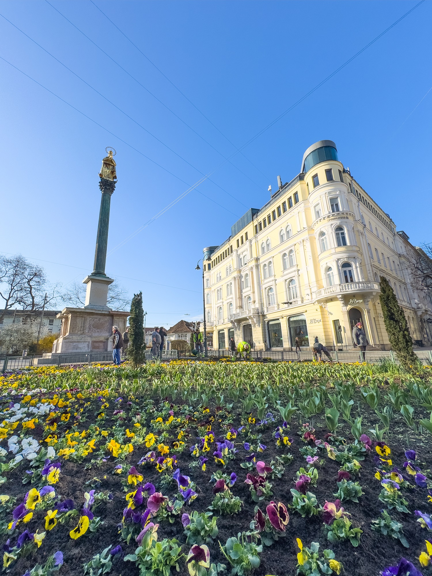 Blick auf ein Blumenbeet am Eisernen Tor in Graz mit einer Statue auf einer Säule im Hintergrund.