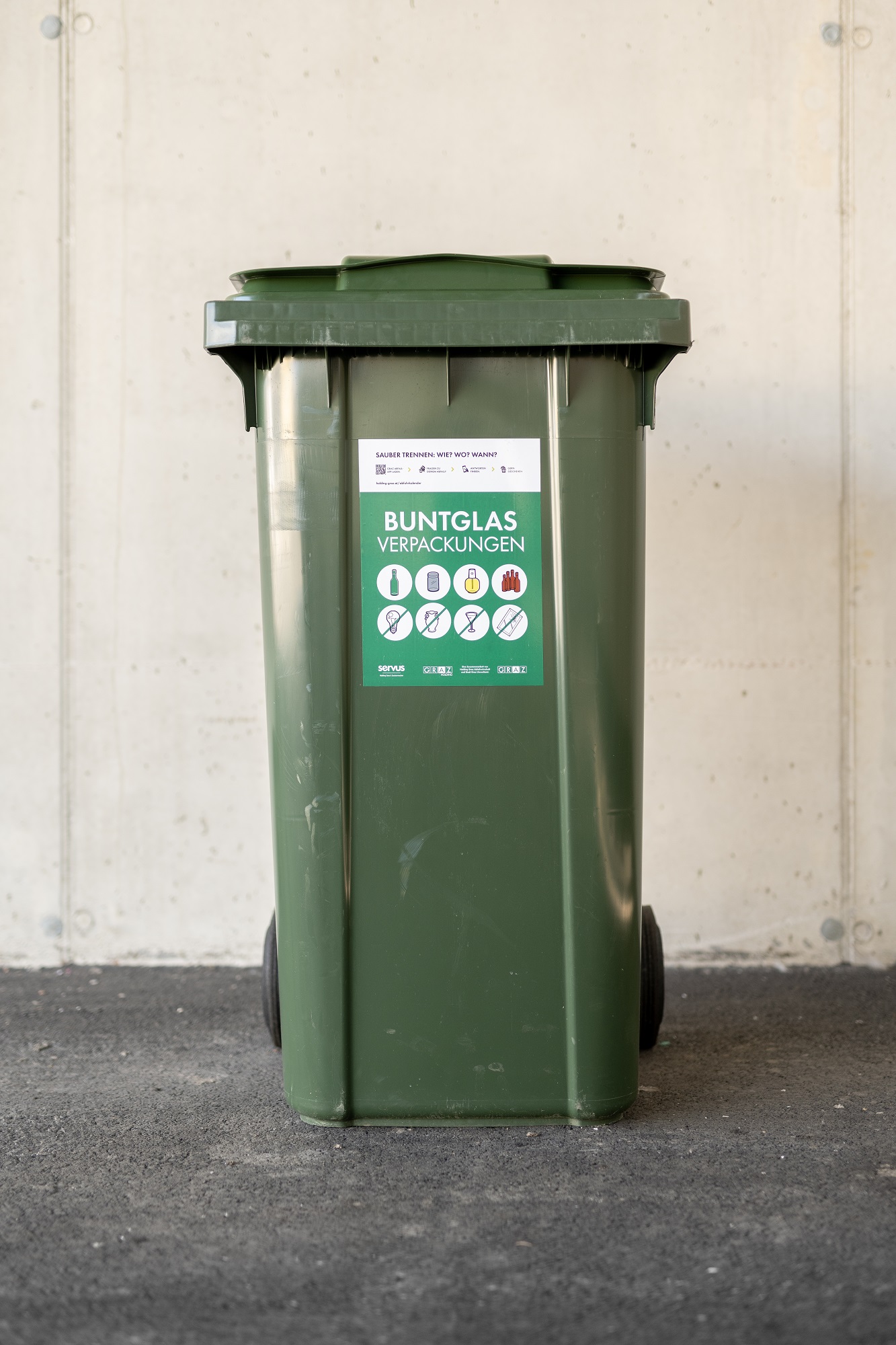 Green recycling bin with a sticker that reads "Buntglas Verpackungen" and has a list of items that can be recycled.