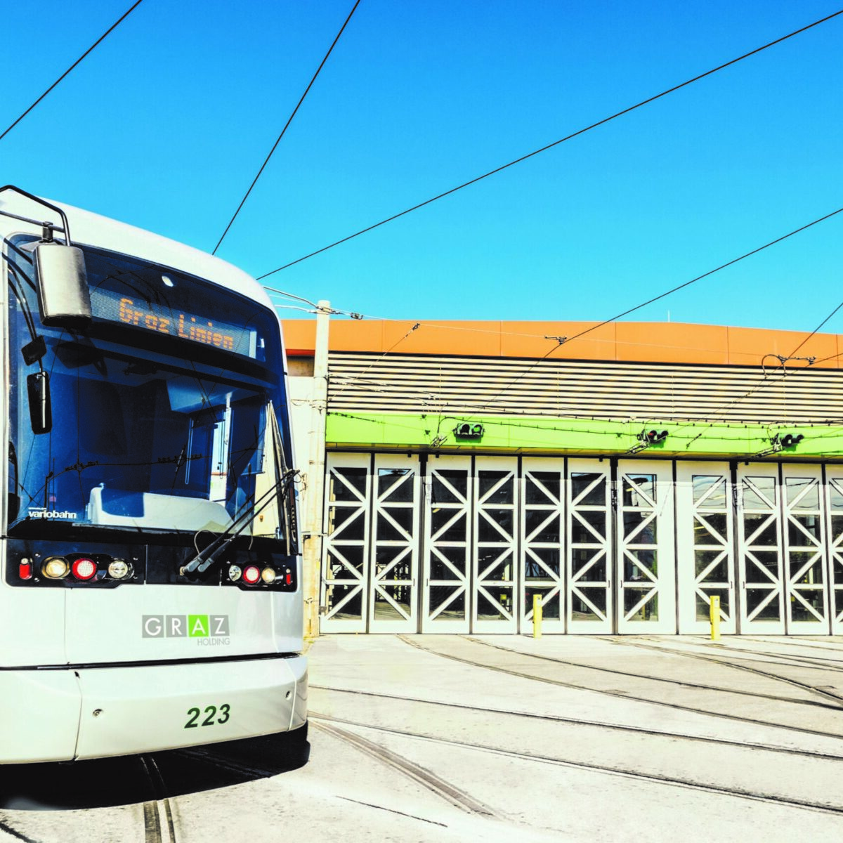 Eine Straßenbahn mit der Nummer 223 und dem Schriftzug "Graz Linien" fährt vor ein Gebäude mit großen, weißen Türen.