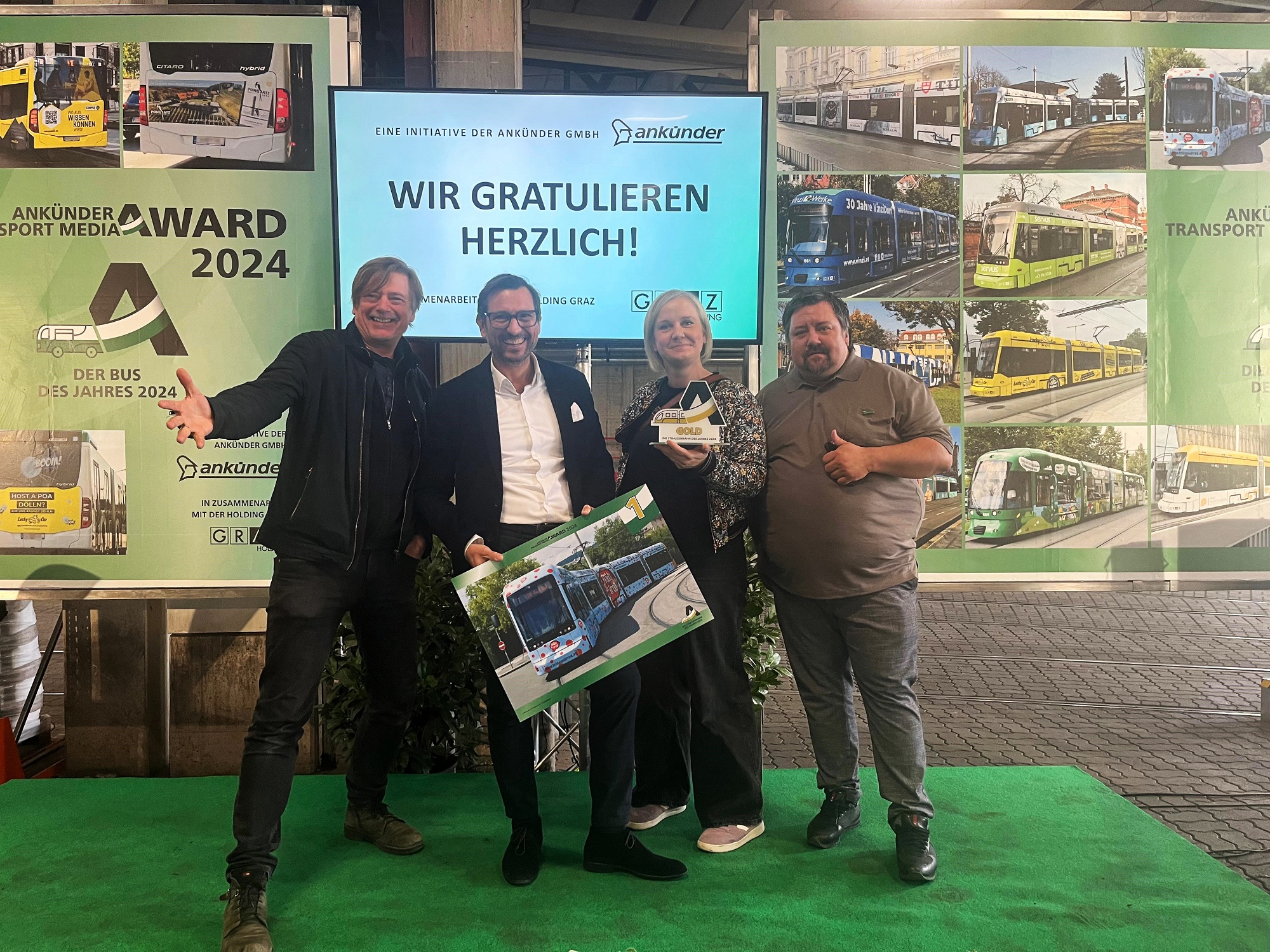 Vier Personen vor einer Wand mit dem Slogan "Wir gratulieren herzlich!" und Bildern von Grazer Straßenbahnen.