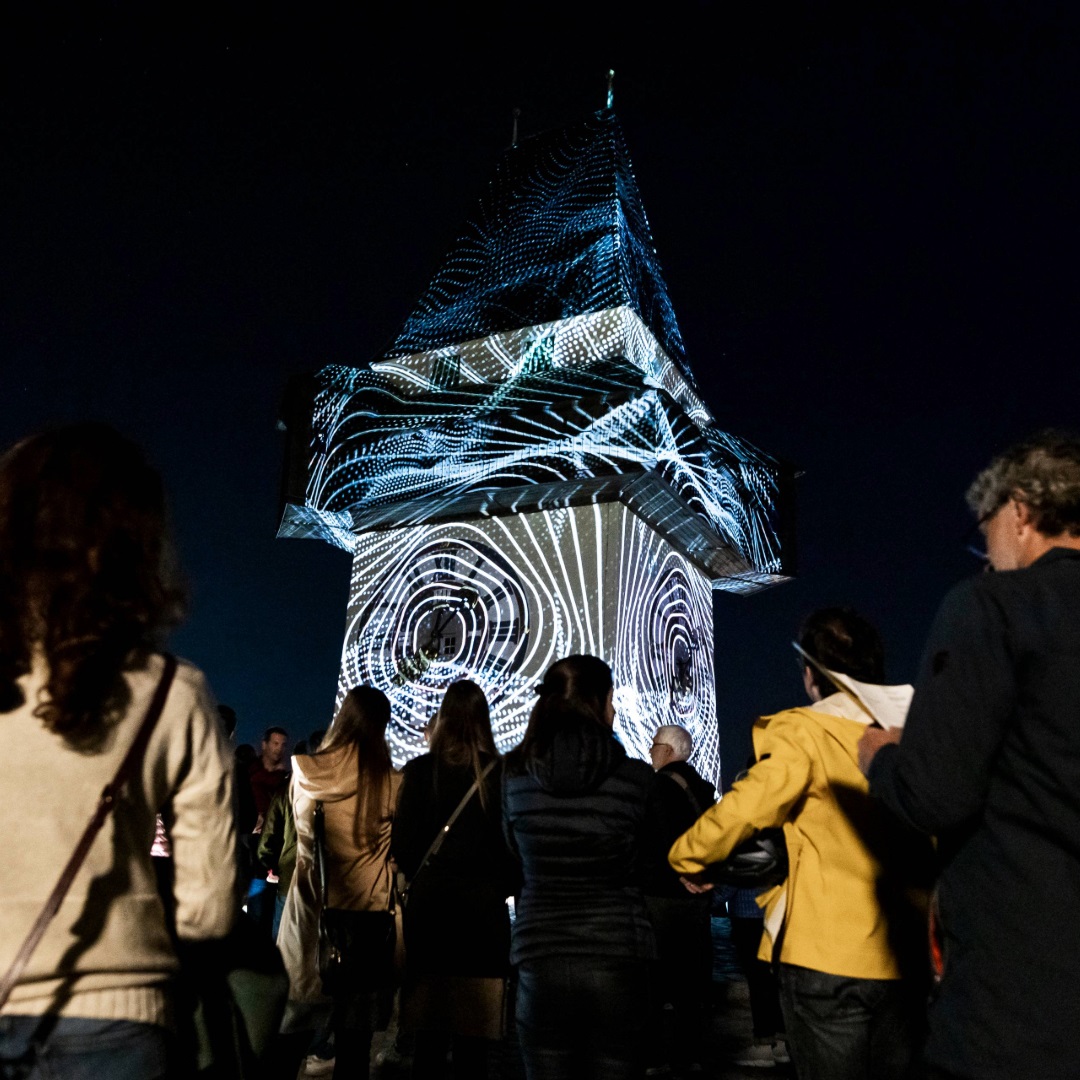 Grazer Uhrturm bei Nacht mit Lichtprojektion aus geometrischen Mustern und Gruppe von Zuschauern.