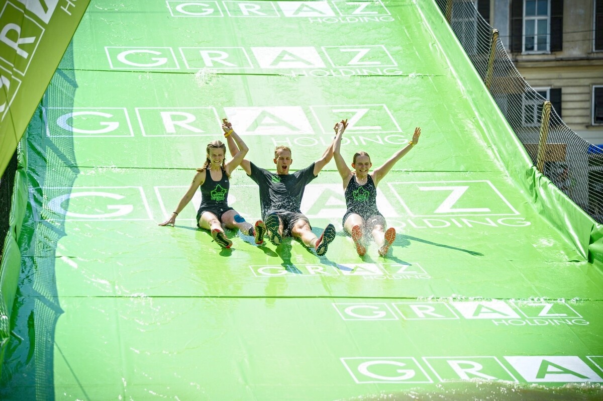 Drei Personen rutschen auf einer grünen Rutsche mit dem Schriftzug "Graz Holding".