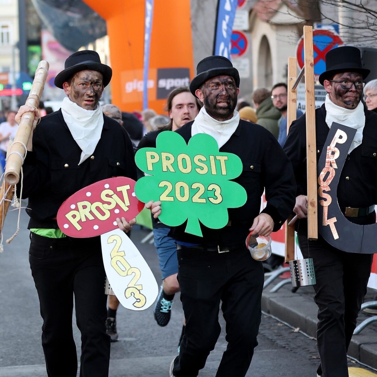 Drei Männer in Rauchfangkehrer-Kostümen laufen mit Schildern „Prosit 2023“ beim Silvesterlauf.