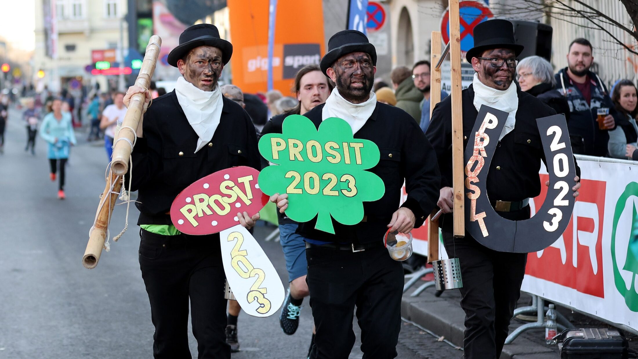 Drei Männer in Rauchfangkehrer-Kostümen laufen mit Schildern „Prosit 2023“ beim Silvesterlauf.