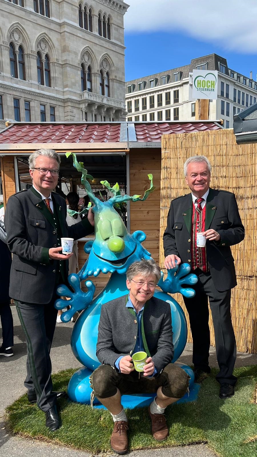 Drei Männer in Tracht posieren mit blauer Fantasiefigur „Wuzzi“, Holzstand und Gebäude im Hintergrund.