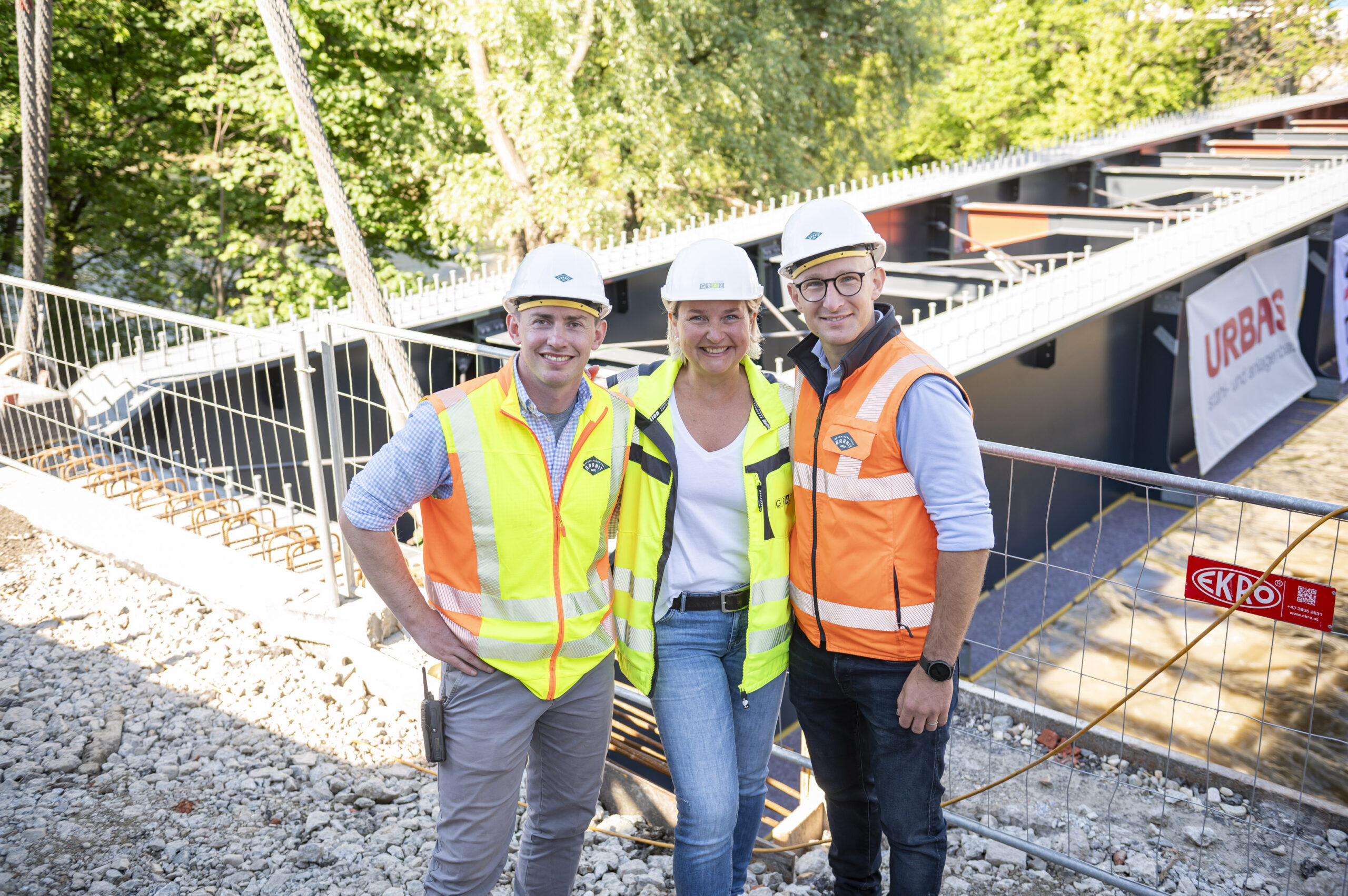 Drei Personen in Sicherheitswesten vor Zaun mit Baustelle dahinter.