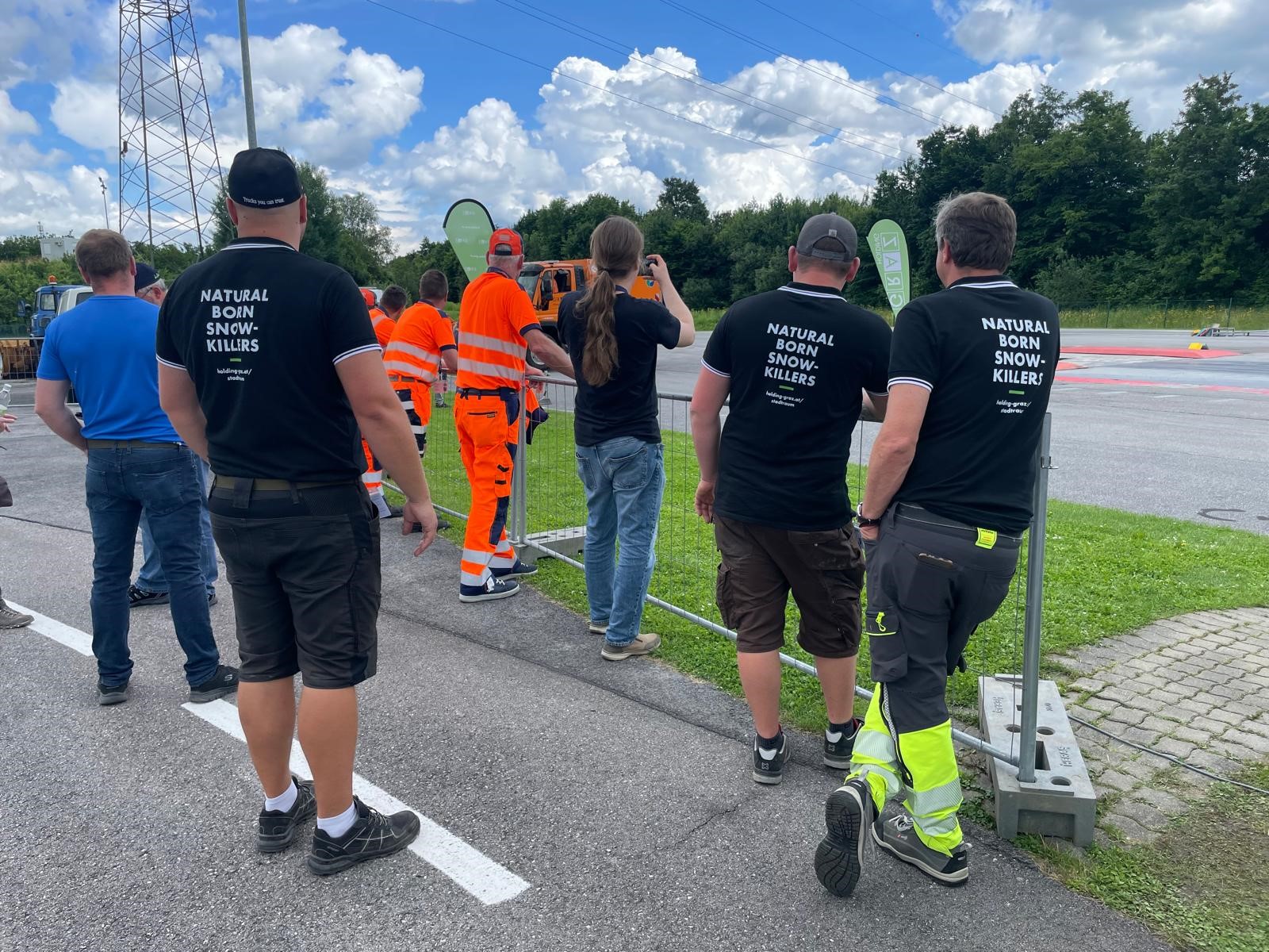 Gruppe von Männern steht vor Zaun und trägt schwarze T-Shirts mit Aufschrift "Natural Born Snow-Killers".