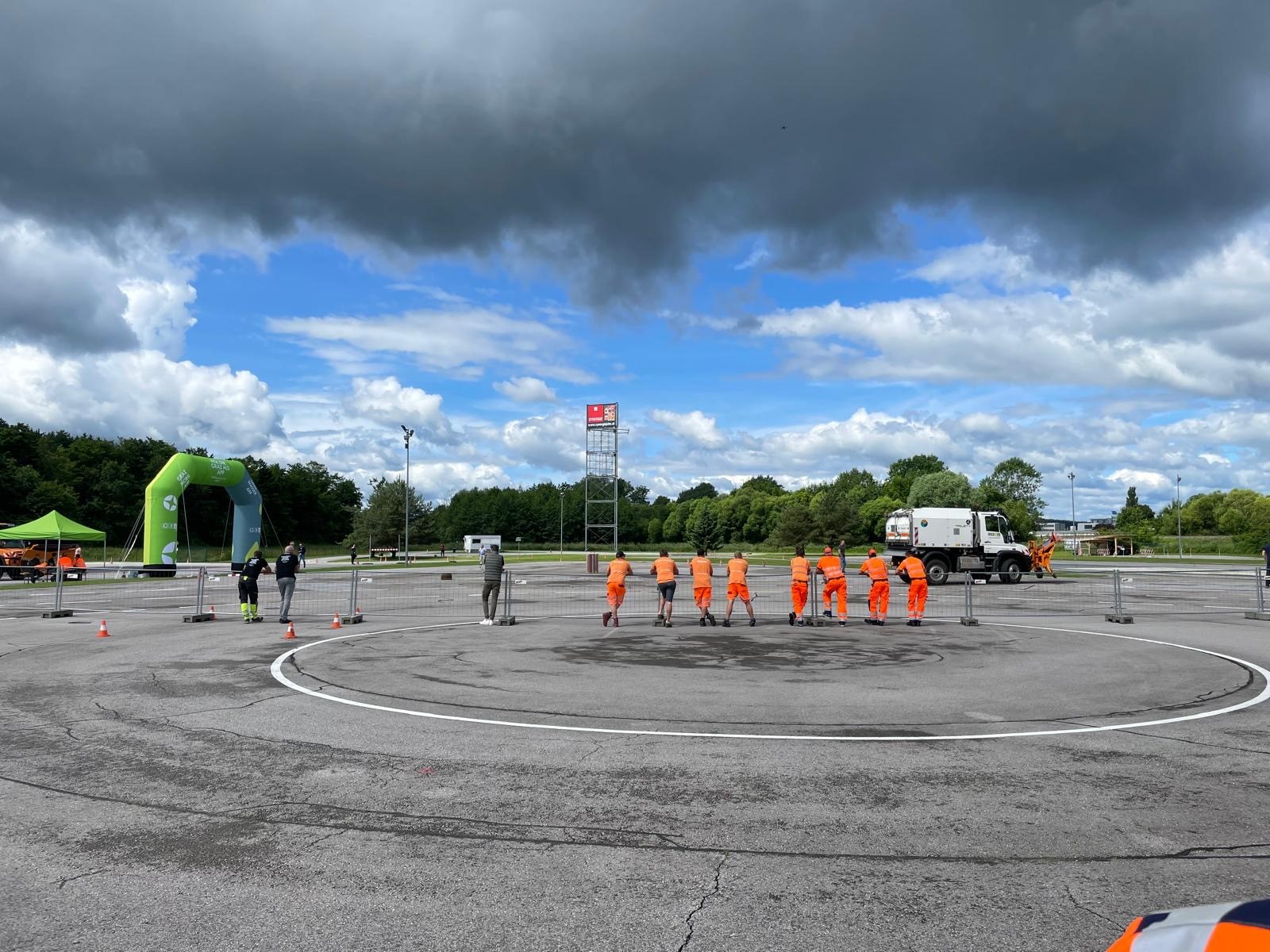 Mehrere Personen lehnen an Zaun auf Rennstrecke und schauen einem Schneepflug zu.