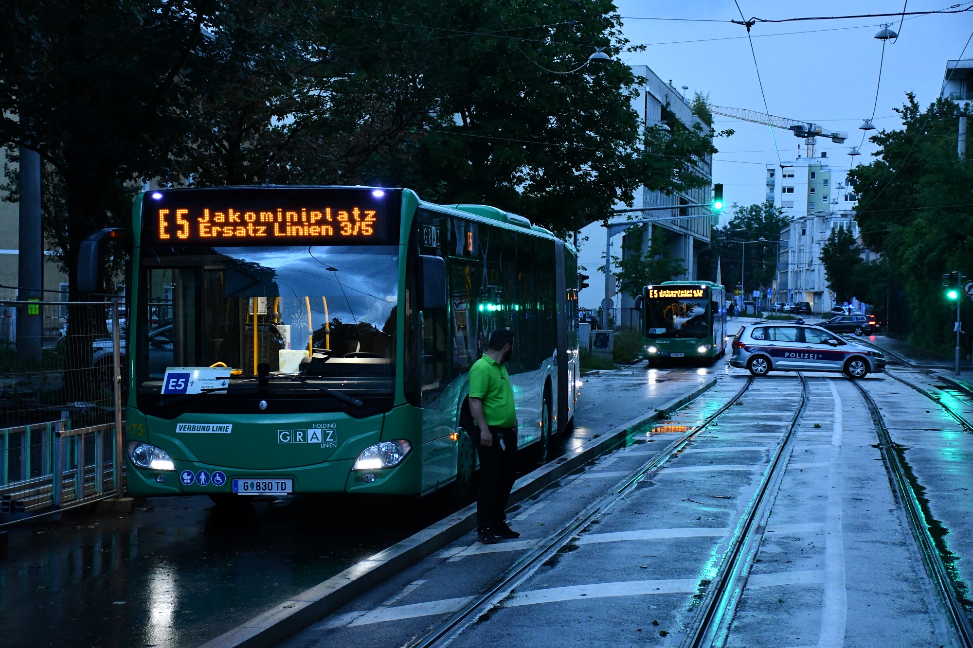 Ersatzbusse der Graz Linien im Regen, Polizei sperrt Straße, nasses Gleisbett reflektiert Lichter.