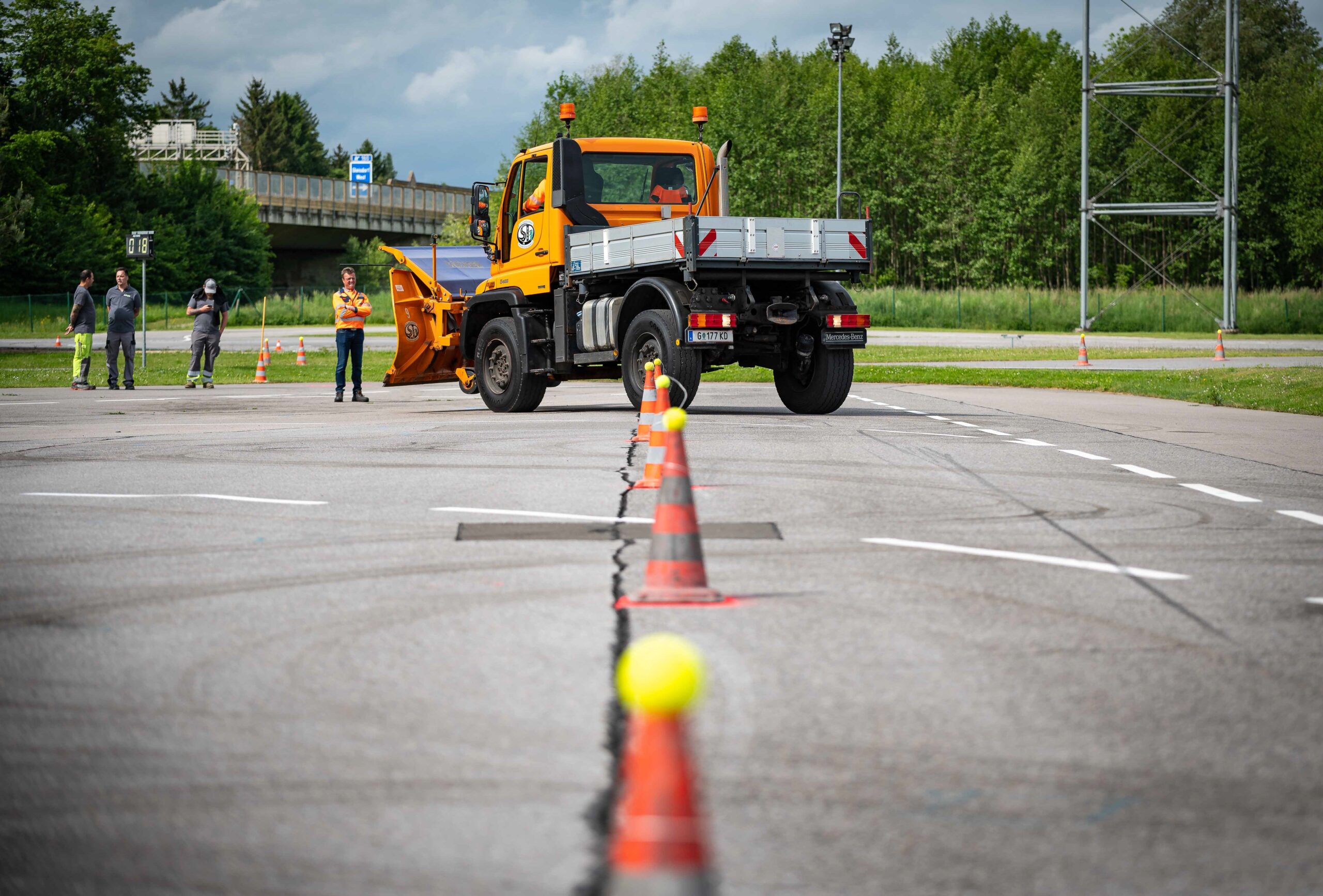 Ein gelber Lastwagen mit Schneepflug steht auf einem Parkplatz. Im Hintergrund sind drei Personen zu sehen.