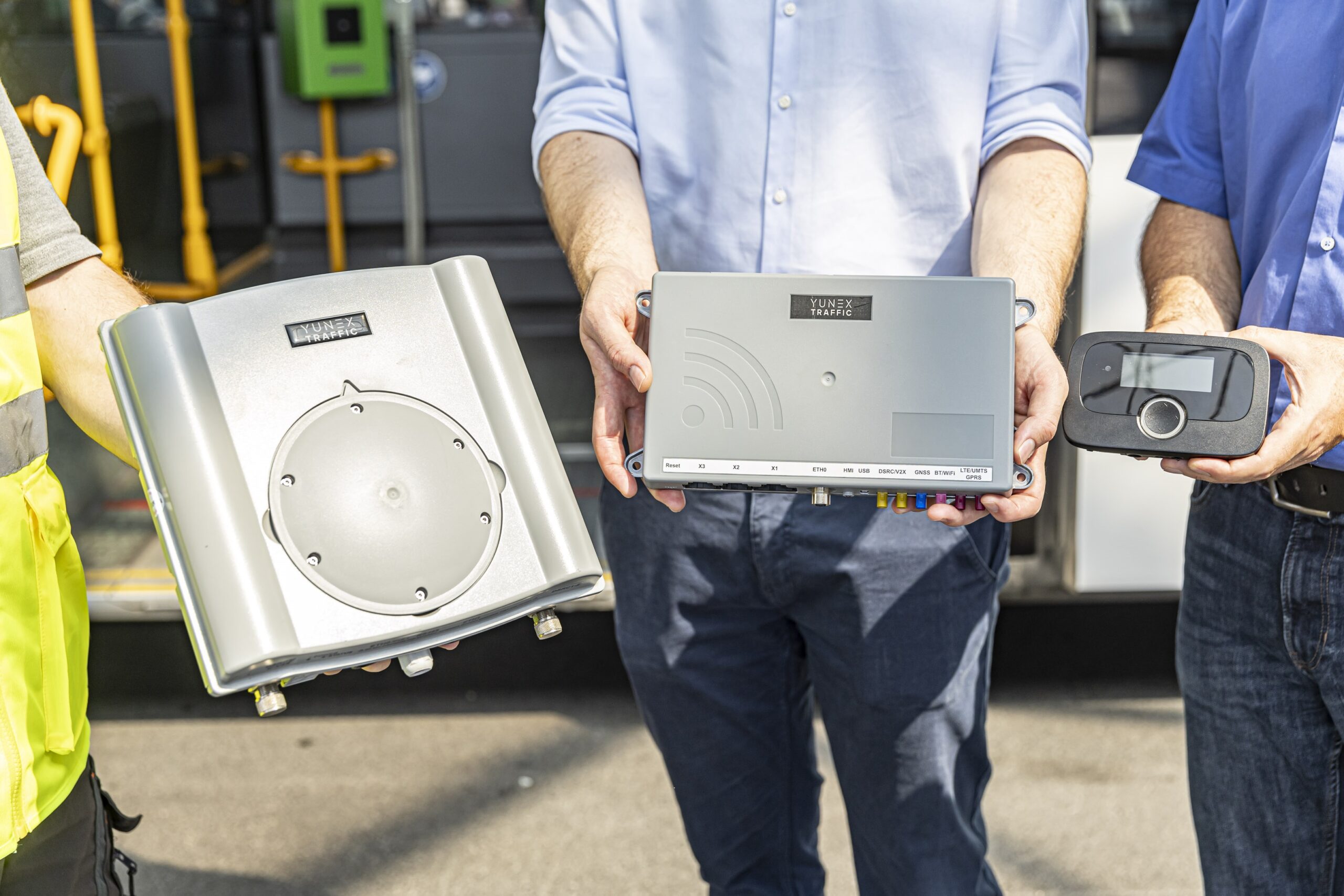 Drei Männer halten „Yunex Traffic“-Geräte vor einem Bus.