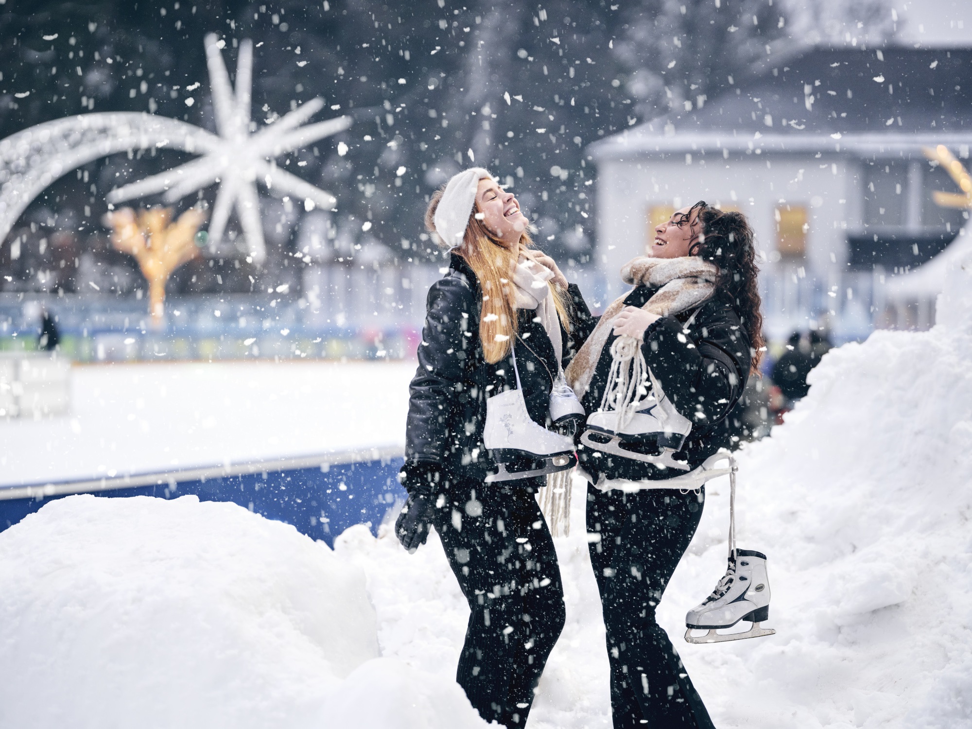 Zwei junge Frauen lachen im Schnee und halten Schlittschuhe in der Hand.