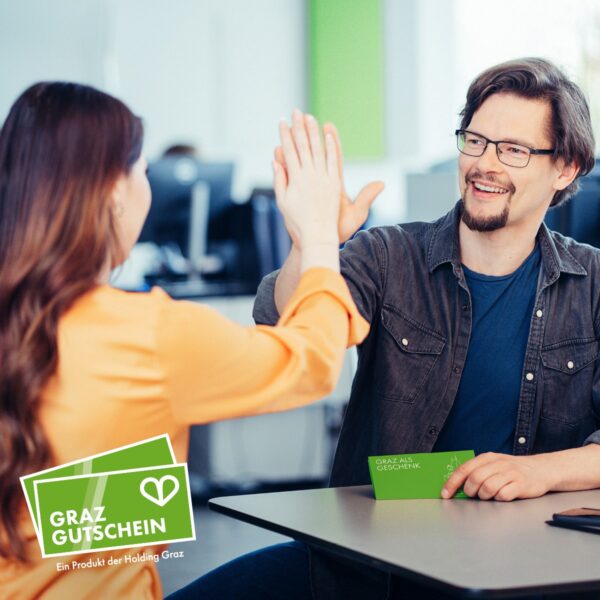 Frau und Mann klatschen ein, Mann hält grünen Umschlag mit Aufschrift "Graz als Geschenk" in Hand.