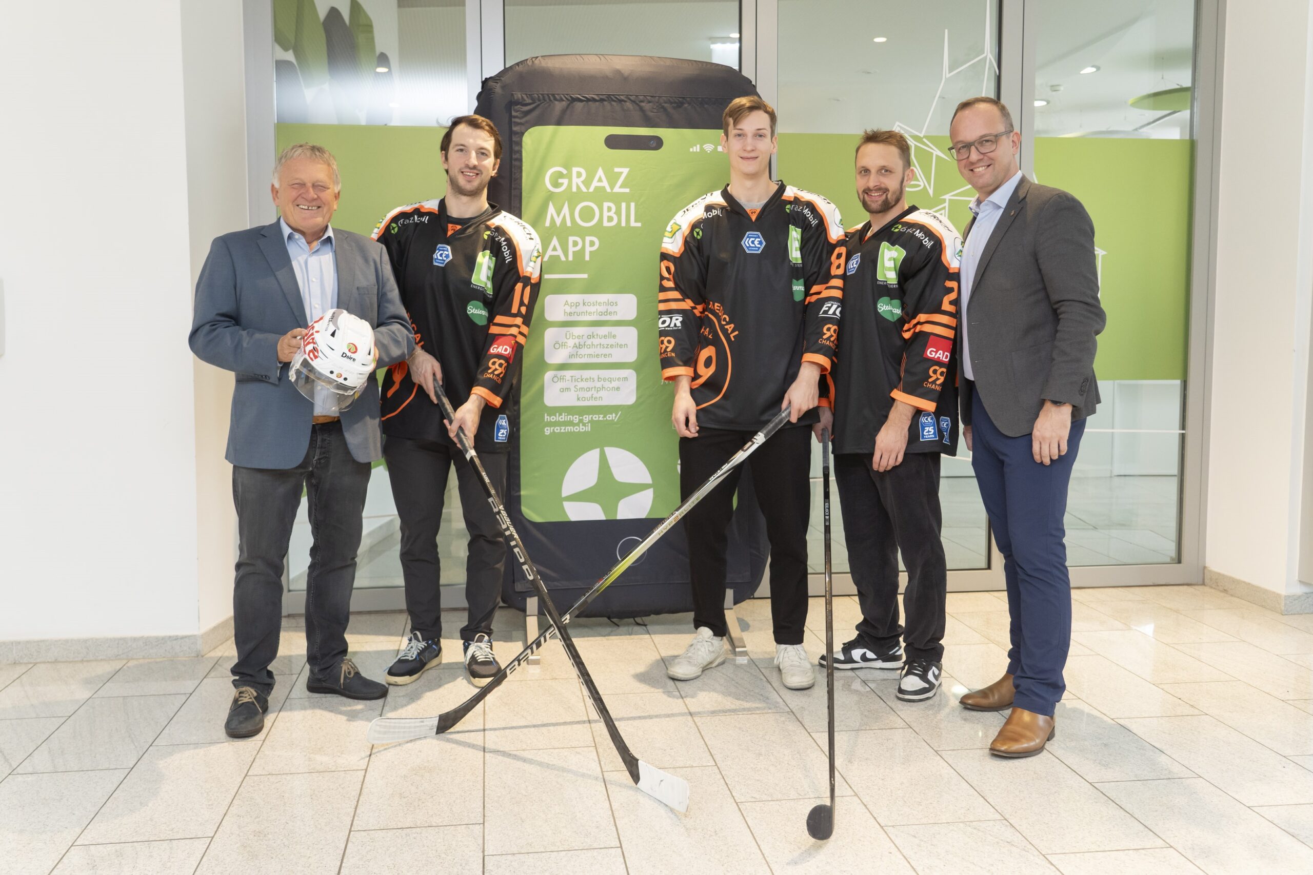 Fünf Männer, darunter Eishockeyspieler, vor großem Smartphone mit Aufschrift "Graz Mobil App".