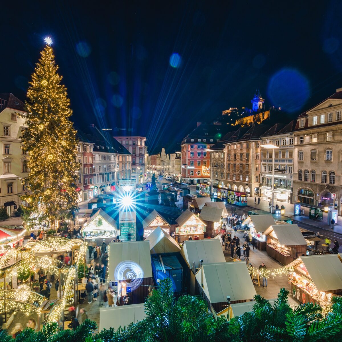 Der Grazer Hauptplatz mit Weihnachtsmarkt und beleuchtetem Christbaum in der Abenddämmerung.