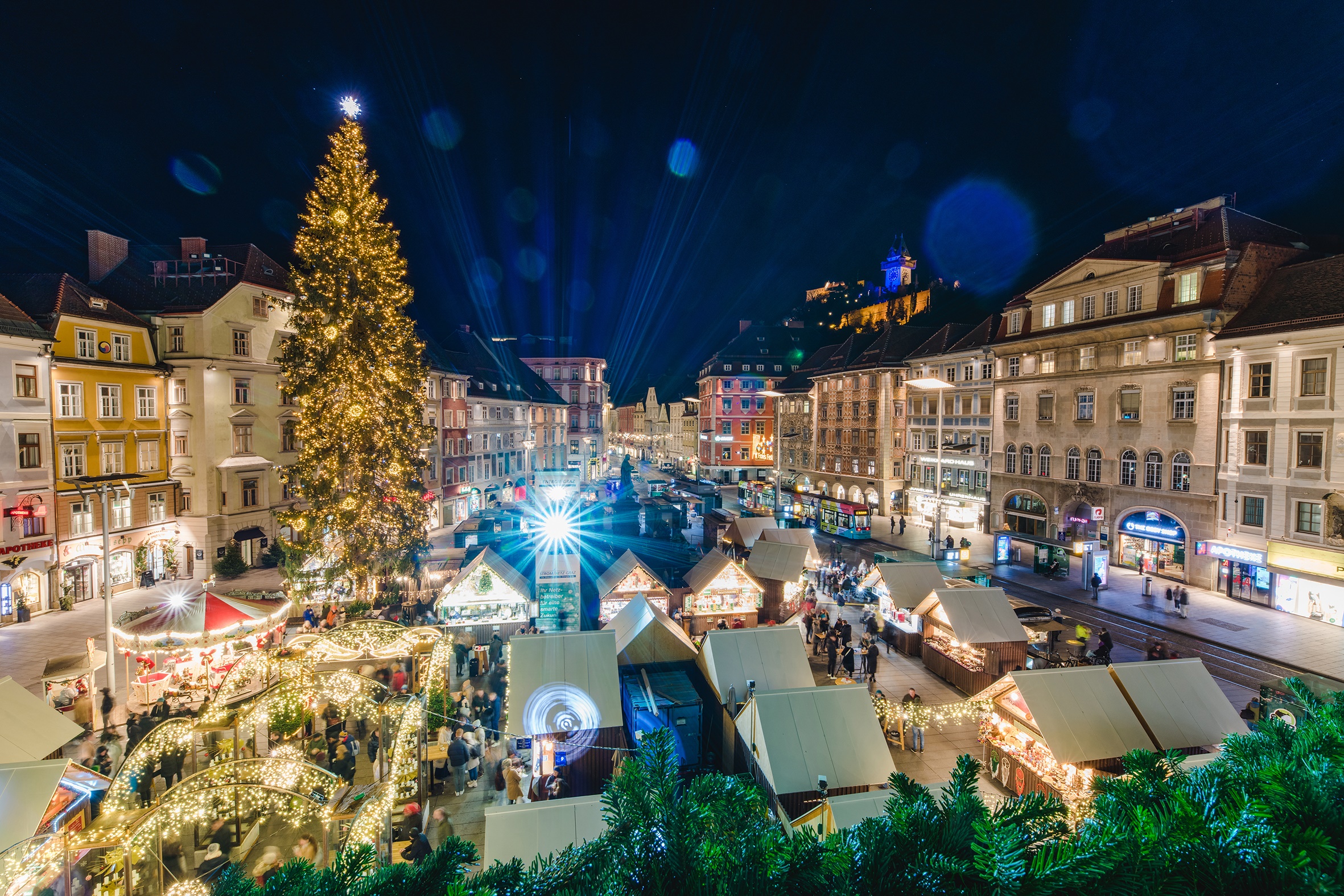 Aerial view of a lit-up Christmas market in Graz at night, with a large Christmas tree in the center
