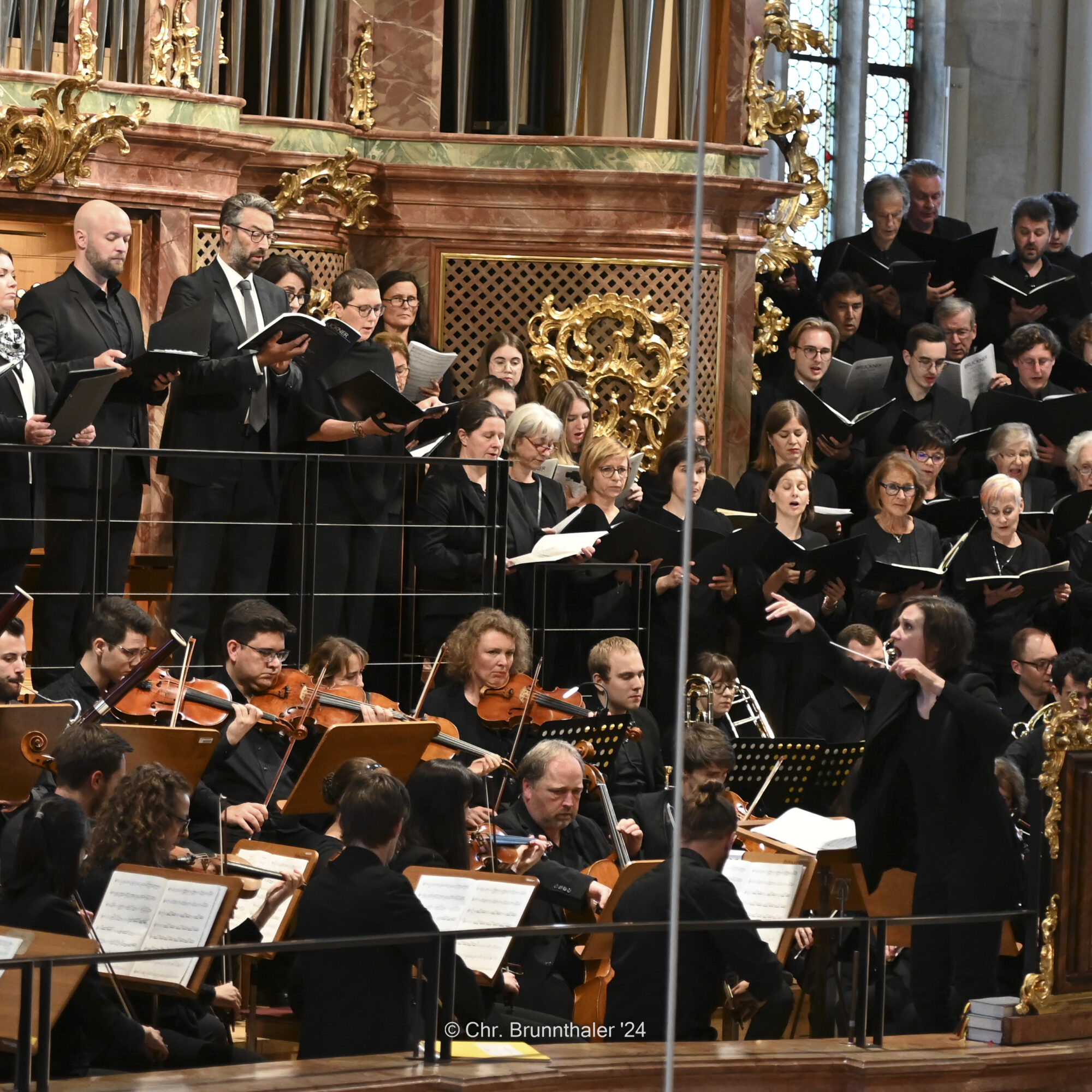 Chor singt auf einer Empore, während Orchester unter Leitung einer Dirigentin spielt.