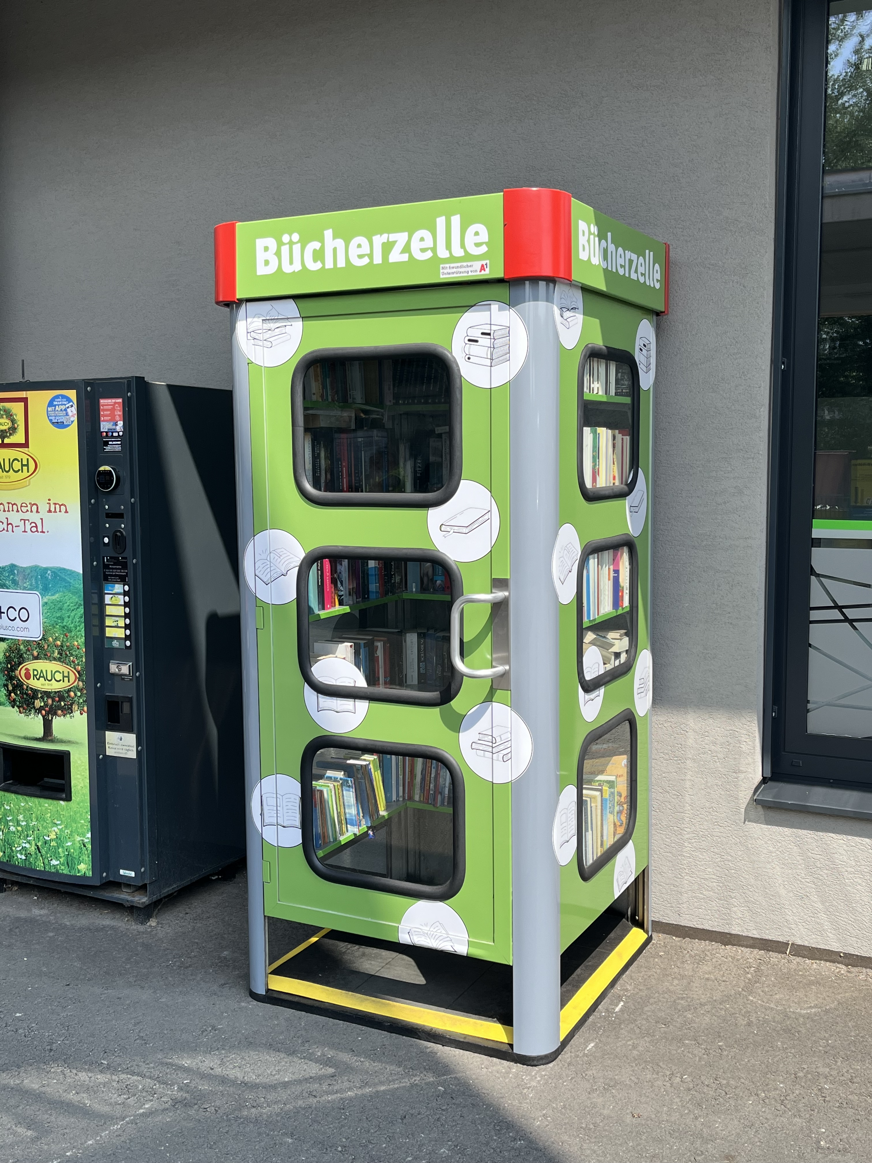 Eine grüne "Bücherzelle" in Graz neben einem Getränkeautomaten.