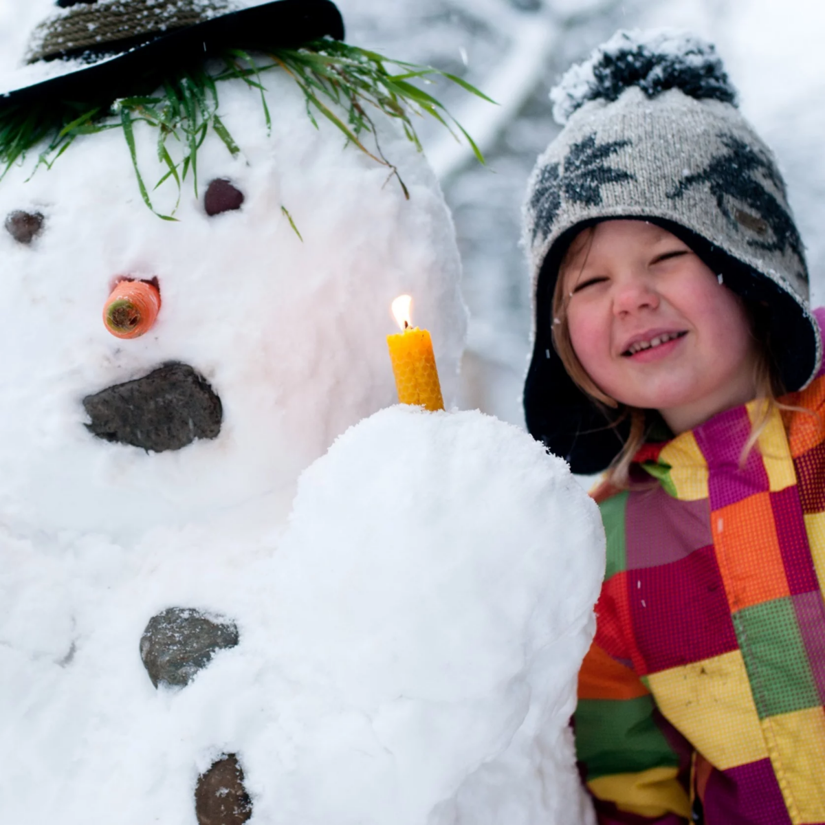 Ein kleines Mädchen hält eine brennende Kerze vor einen Schneemann.
