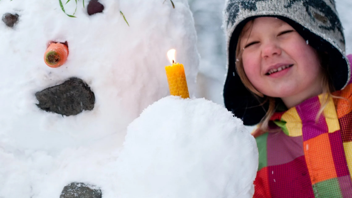 Ein kleines Mädchen hält eine brennende Kerze vor einen Schneemann.