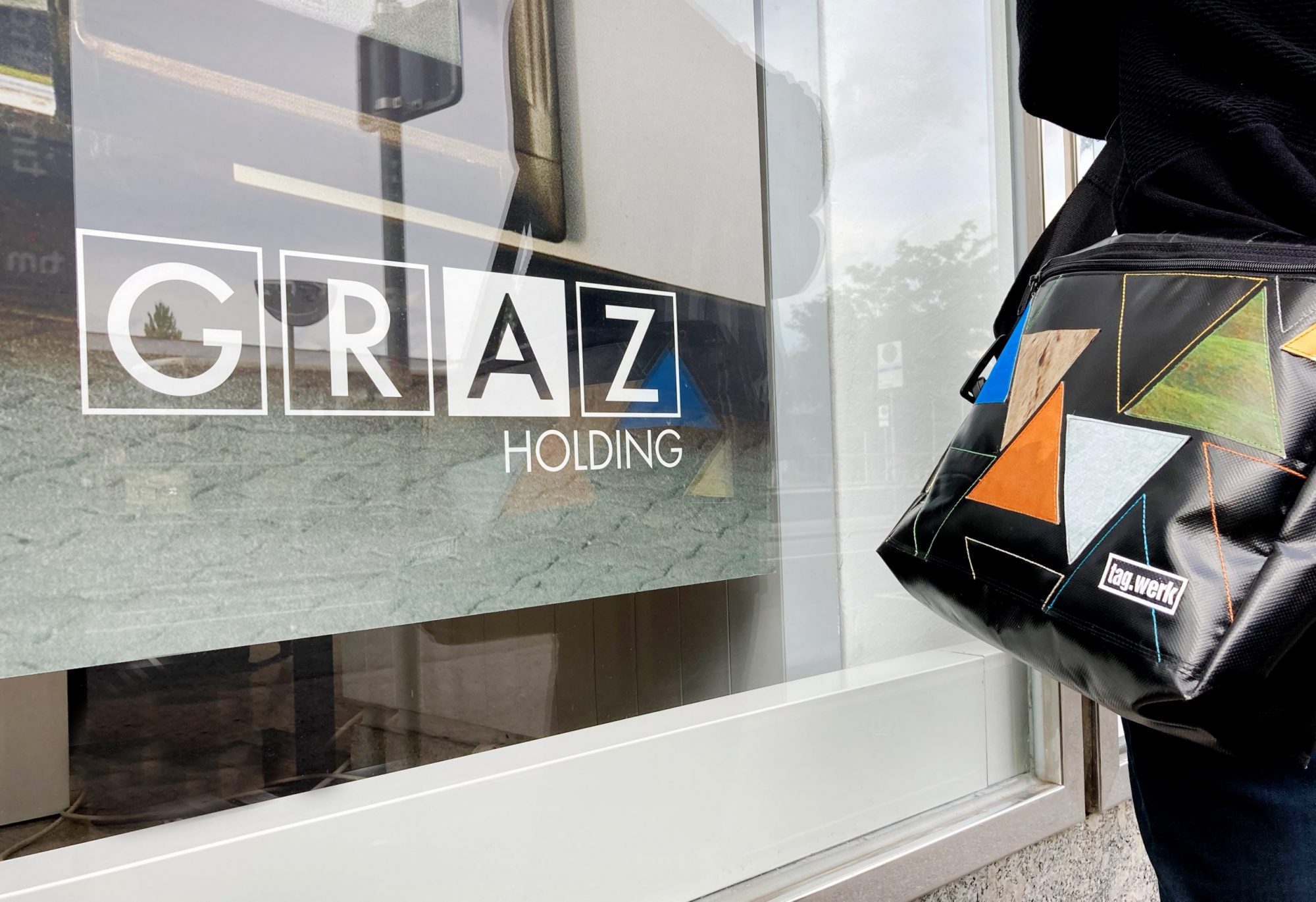 Schwarze tag.werk-Tasche vor einem Fenster mit dem Schriftzug "Graz Holding"