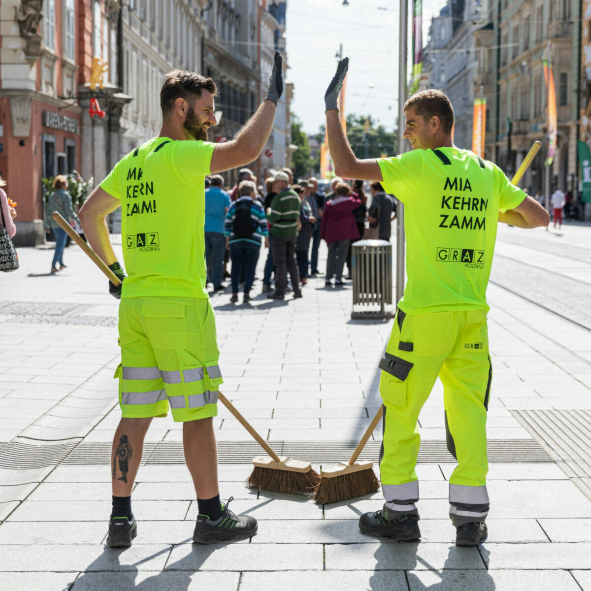Zwei Straßenreiniger in neongelben Uniformen geben sich ein High-Five in belebter Fußgängerzone.