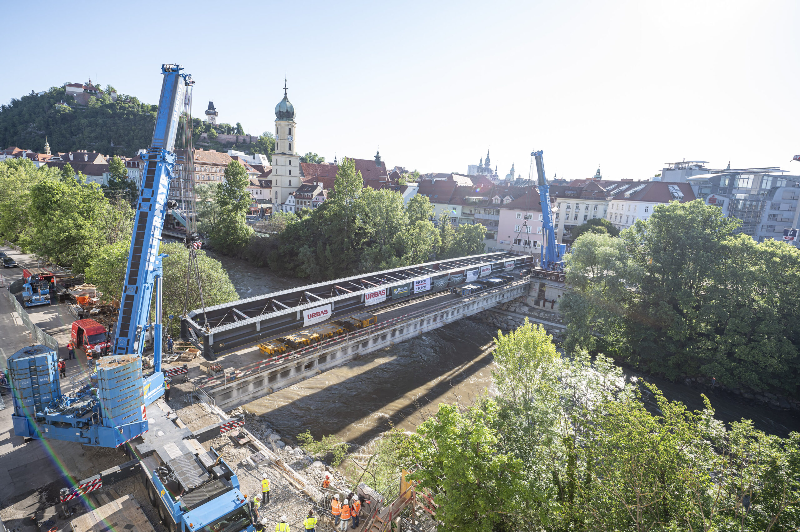 Zwei Kräne heben ein massives Brückenteil über die Mur in Graz.