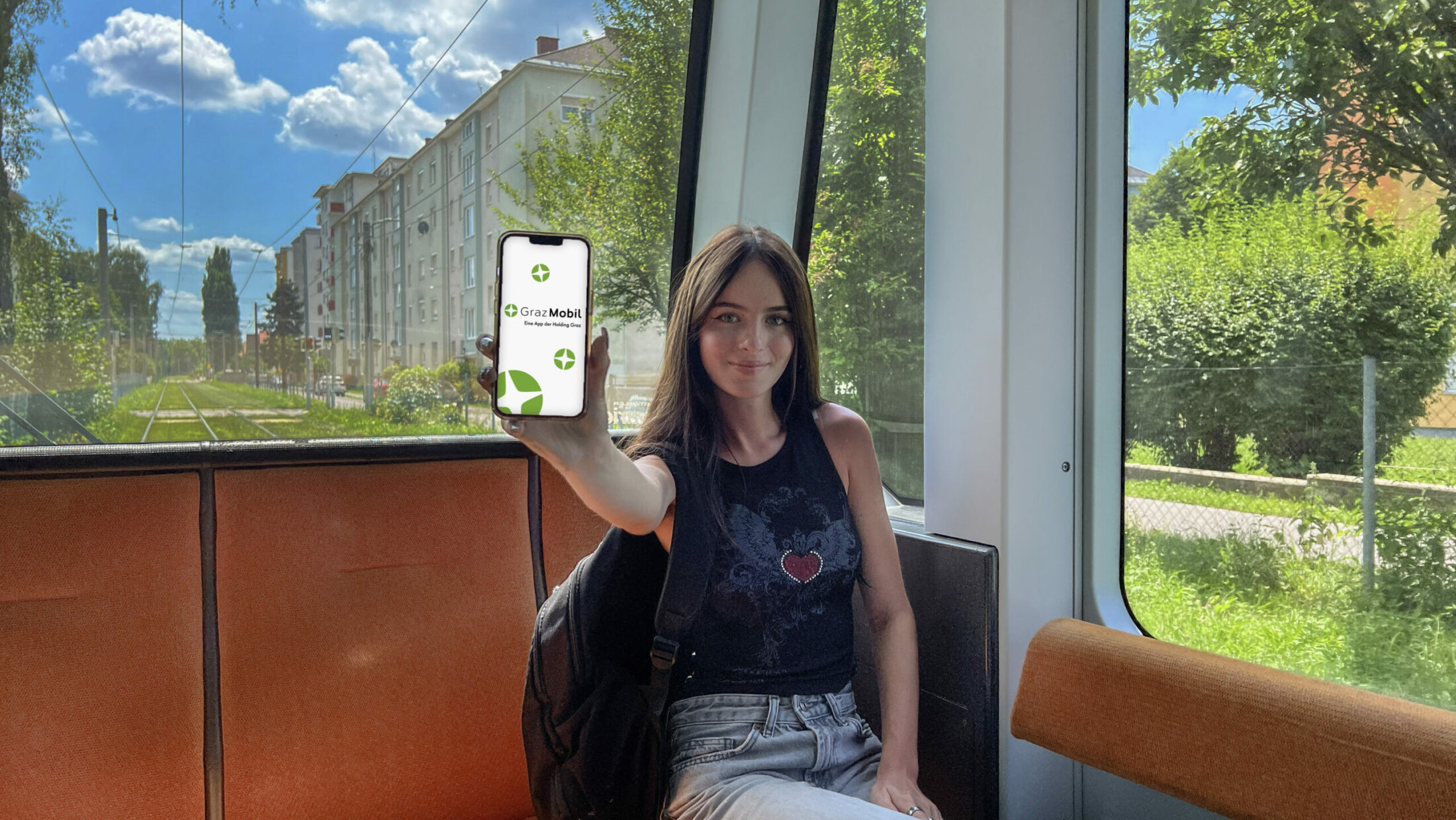 Junge Frau sitzt in Straßenbahn und hält Smartphone mit geöffneter "GrazMobil"-App in Hand.