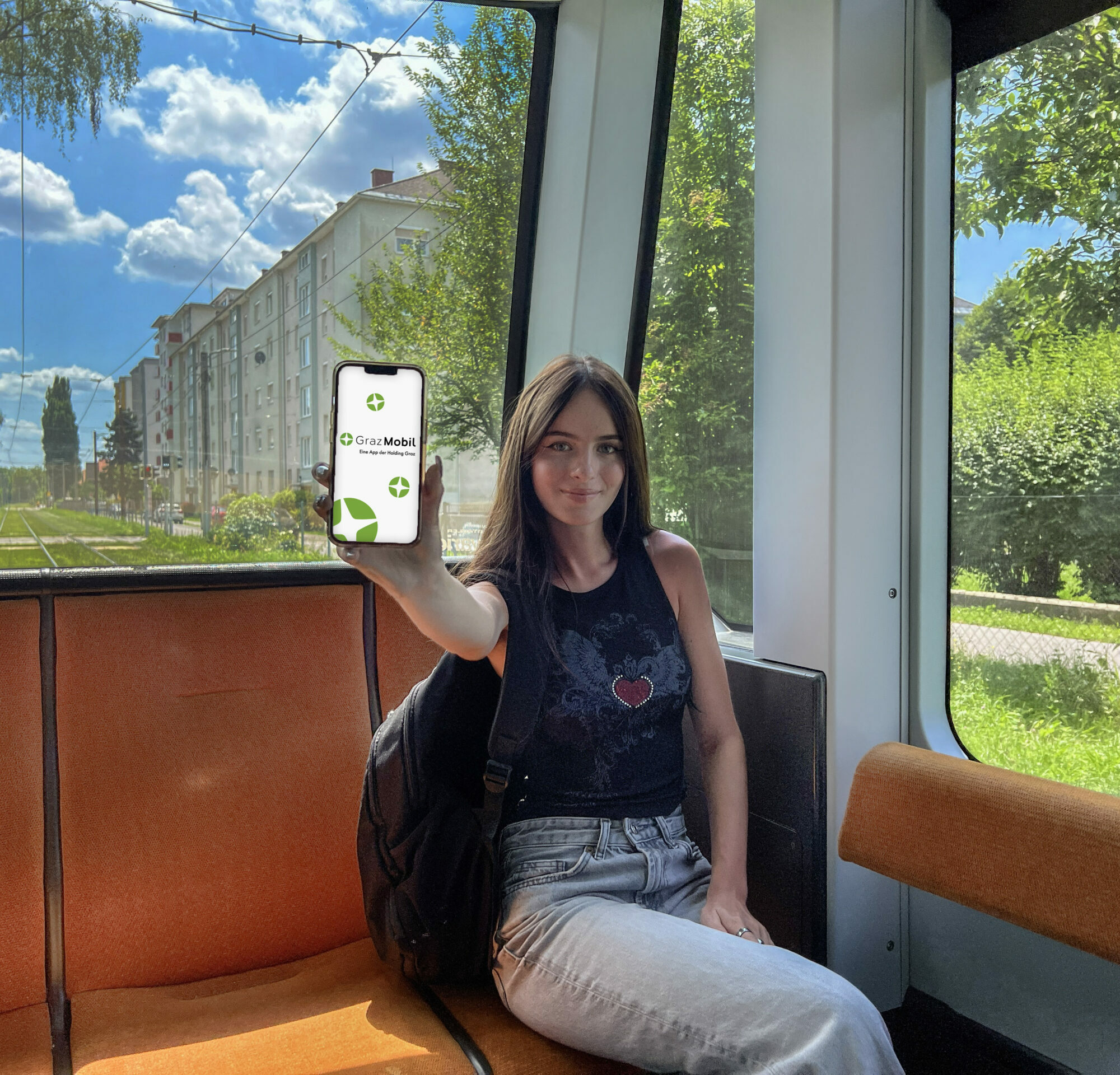 Teenager, weiblich, sitzt in der hintersten Reihe einer Straßenbahn. Sie hält ein Handydisplay in der Hand, welches die GrazMobil-App zeigt