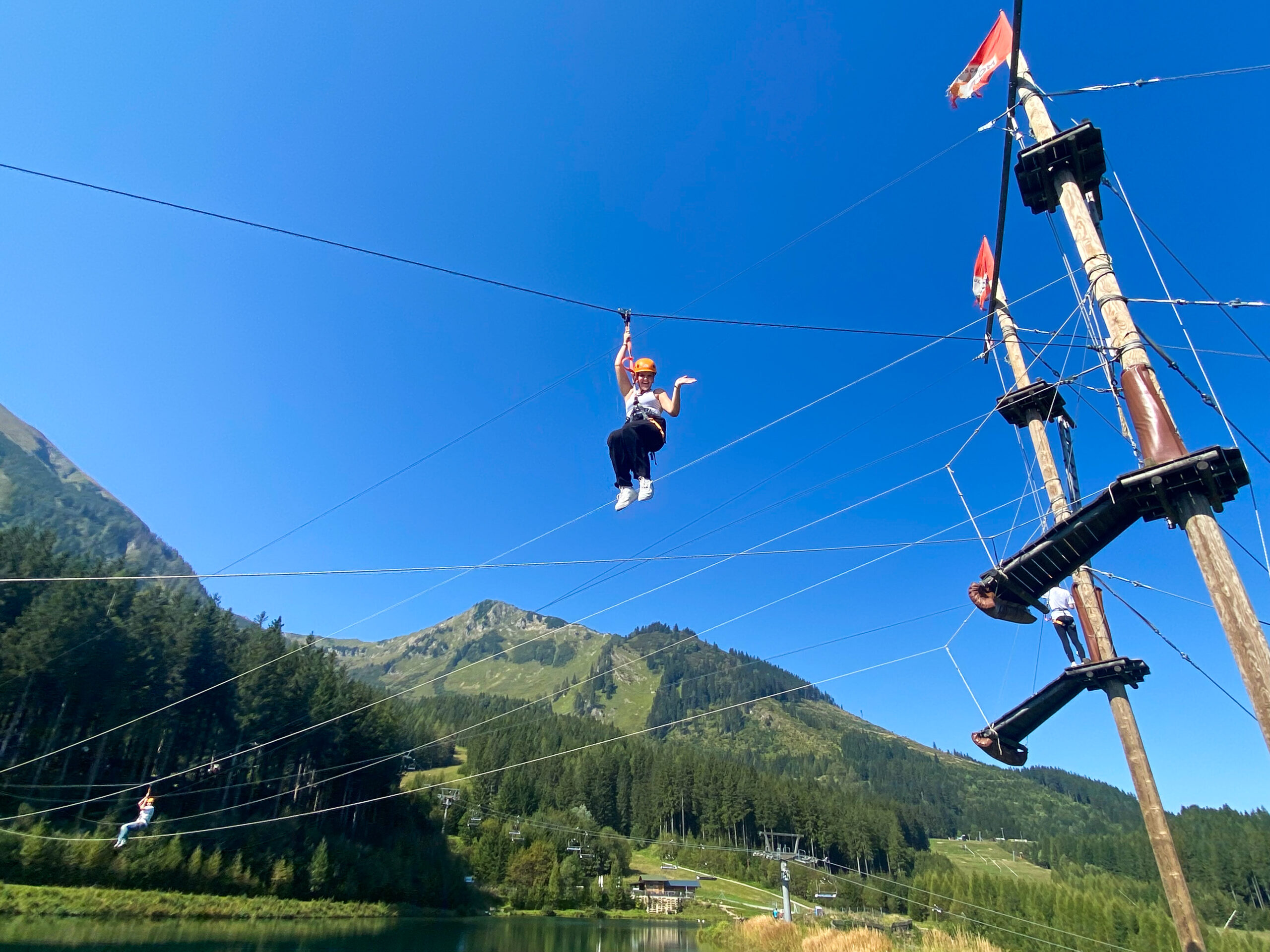 Eine Frau schwebt in alpiner Umgebung an einem Seil über einem See und einem Wald.