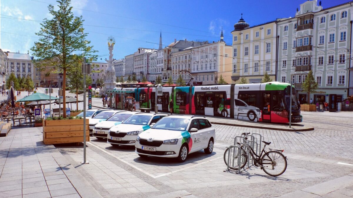 Vier tim-Fahrzeuge am am Linzer Hauptplatz, dahinter fährt mit tim gebrandete Straßenbahn vorbei.
