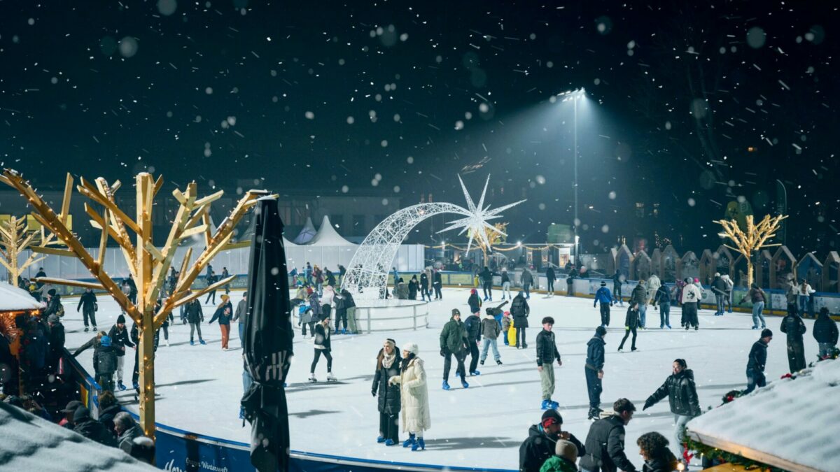 Nighttime view of an outdoor ice rink with people skating and snow falling. "Have a nice ice day!" is written on the side of the rink.