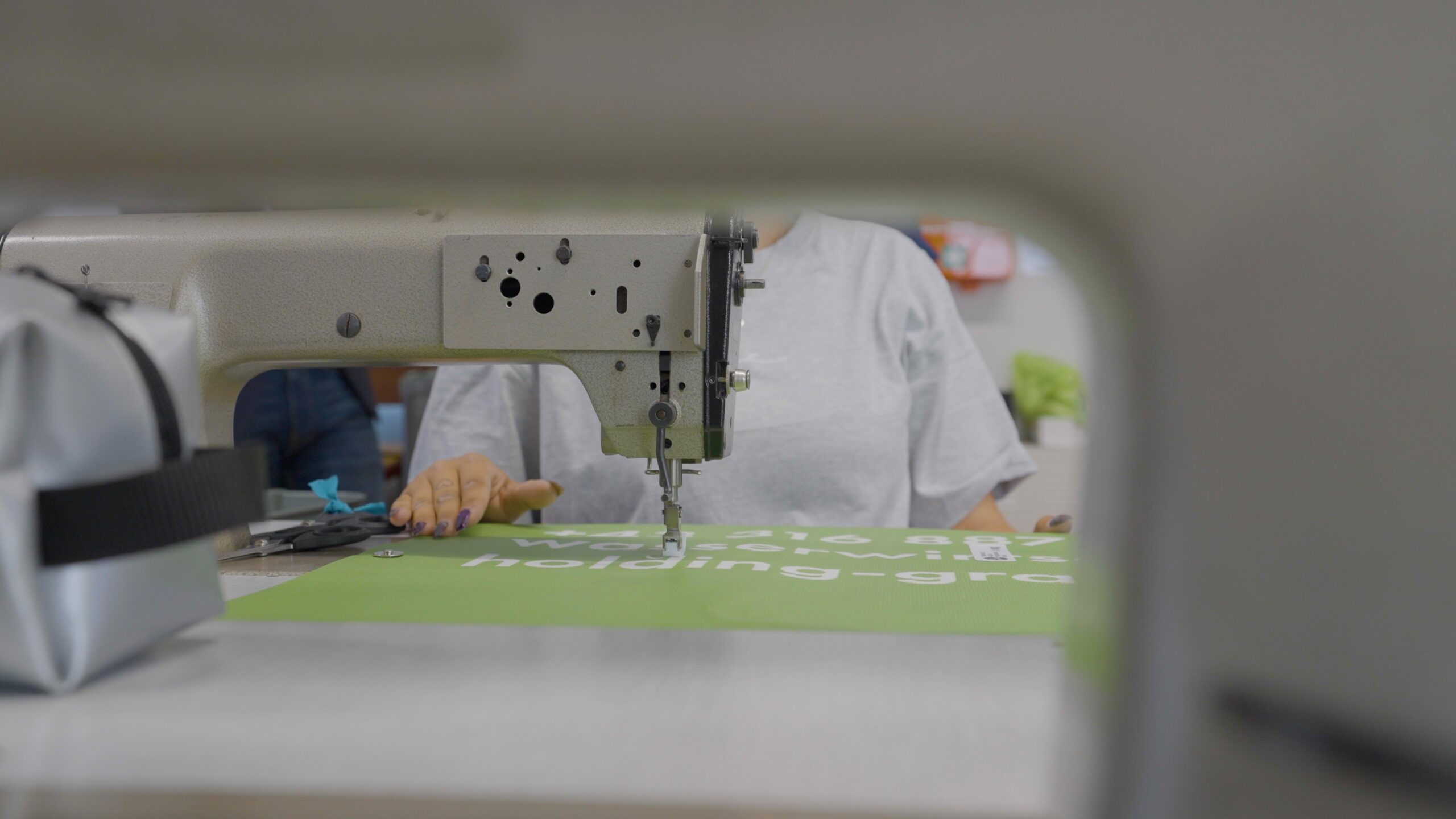 Frau näht an Nähmaschine, Vordergrund unscharf, grüne Stoffbahn mit weißer Schrift sichtbar.