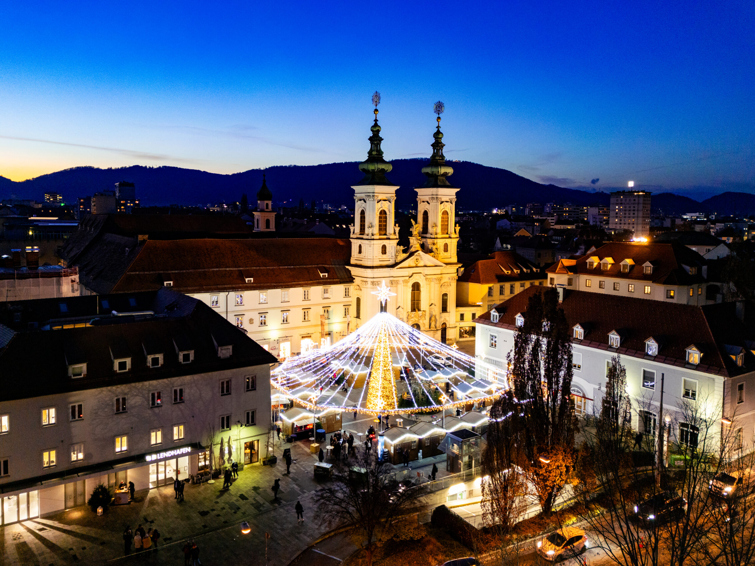 Aerial view of Graz's Christmas market at dusk, with church backdrop.