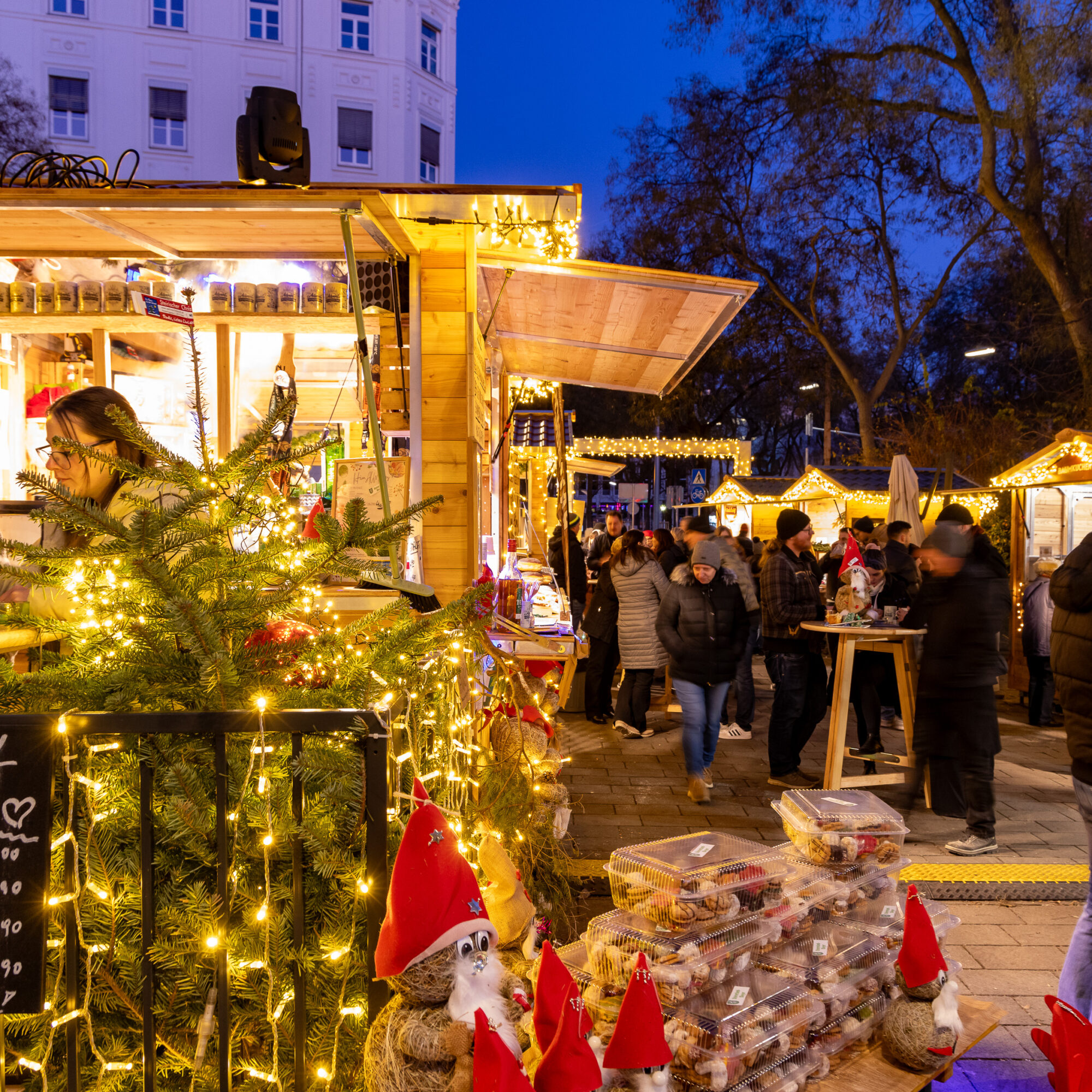 Weihnachtsmarkt in Graz. Helle Holzstände, Lichterketten, Publikum. „Wichtl sucht zu Hause“-Schild.