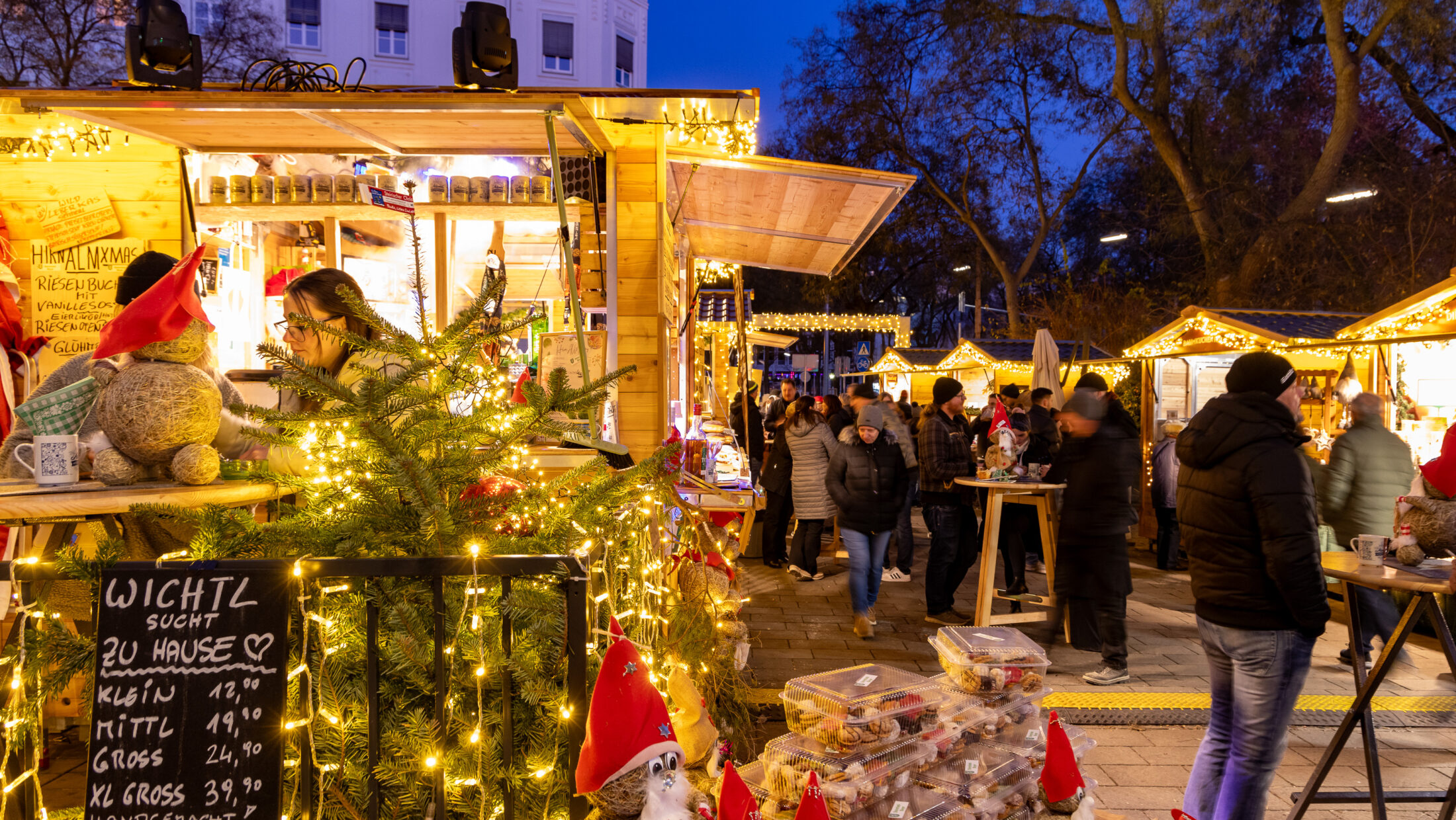 Weihnachtsmarkt in Graz. Helle Holzstände, Lichterketten, Publikum. „Wichtl sucht zu Hause“-Schild.