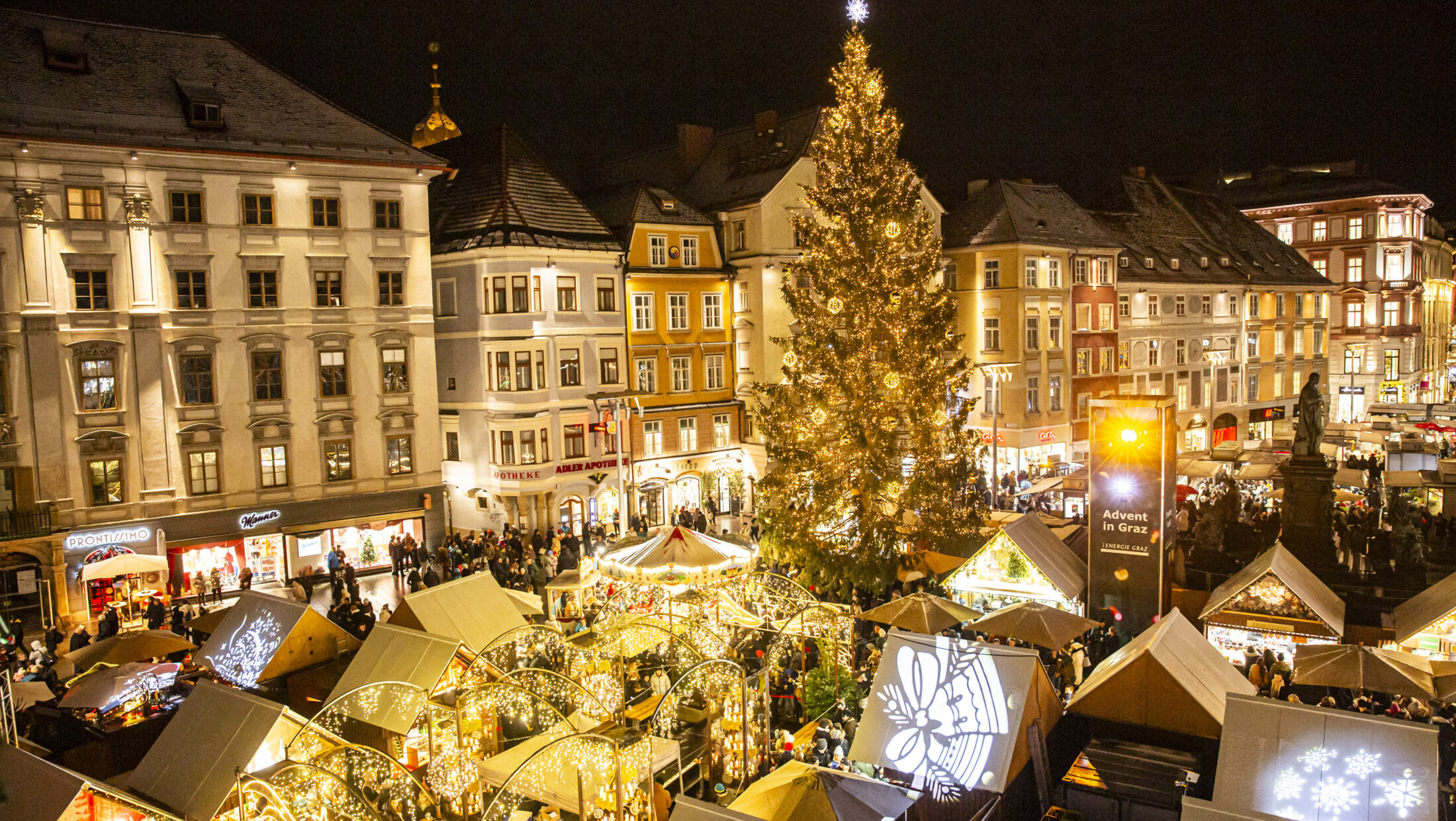 Graz Christmas market at night with illuminated stalls and large Christmas tree.