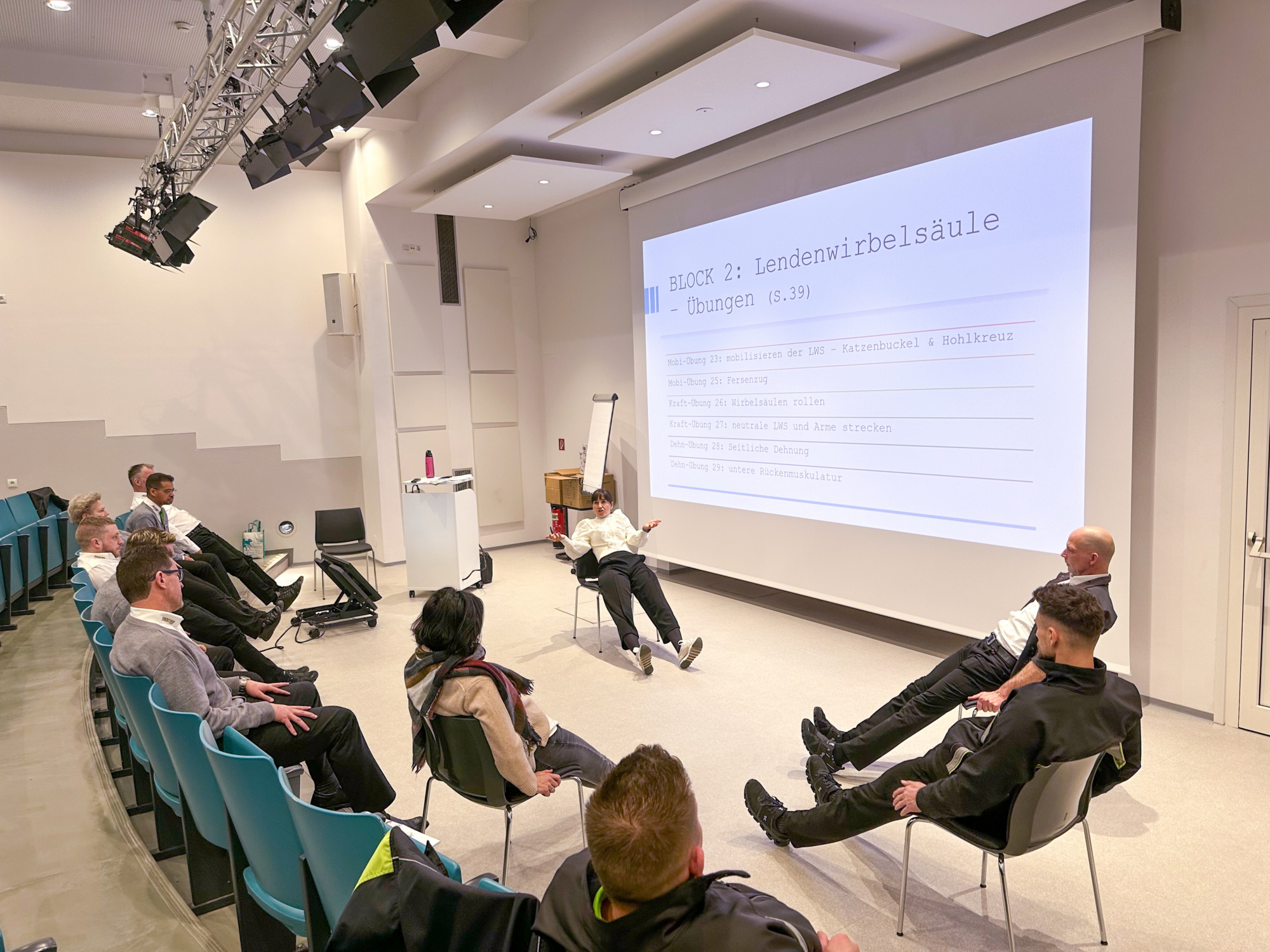 Eine Gruppe von Menschen sitzt in einem Hörsaal und macht Übungen für die Lendenwirbelsäule.
