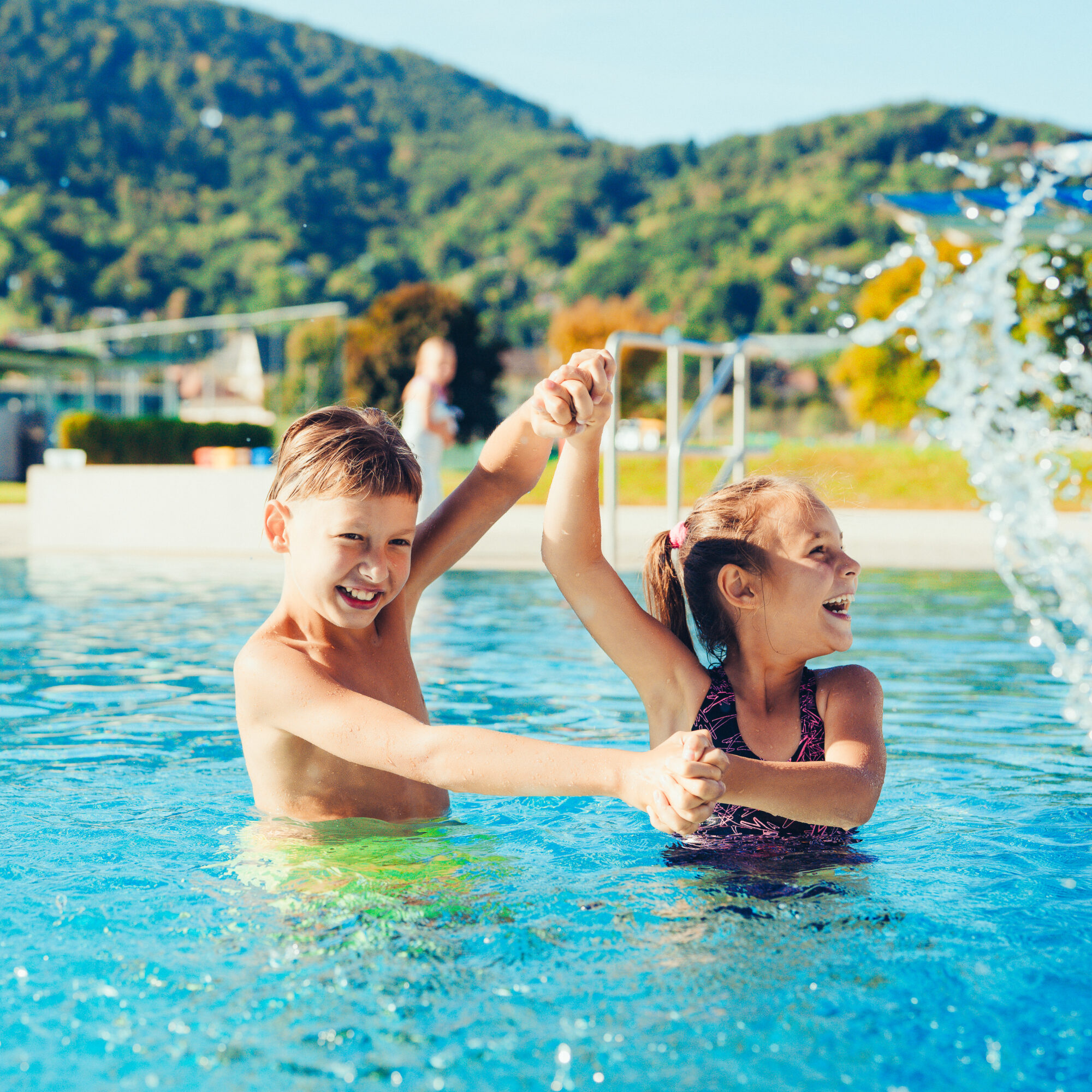 Zwei fröhliche Kinder plantschen im Auster Freibad Graz mit Bergkulisse.
