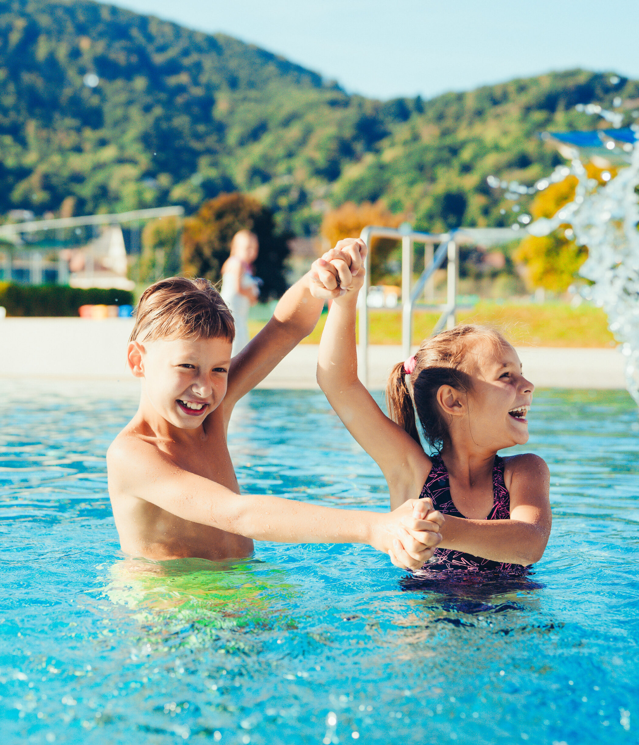 Zwei fröhliche Kinder plantschen im Auster Freibad Graz mit Bergkulisse.